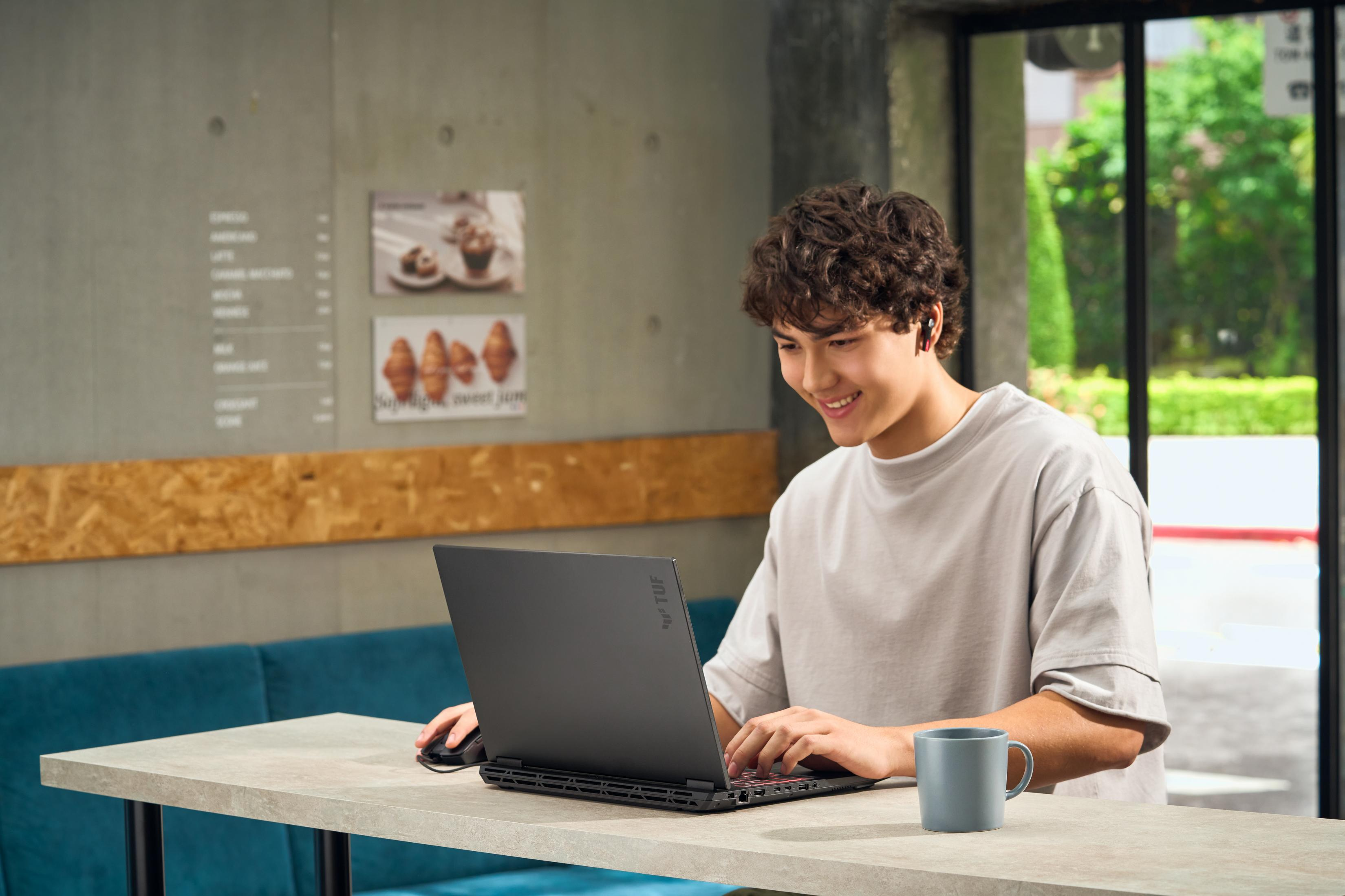 A young man with curly hair types on a black laptop, with a blue mug beside him.