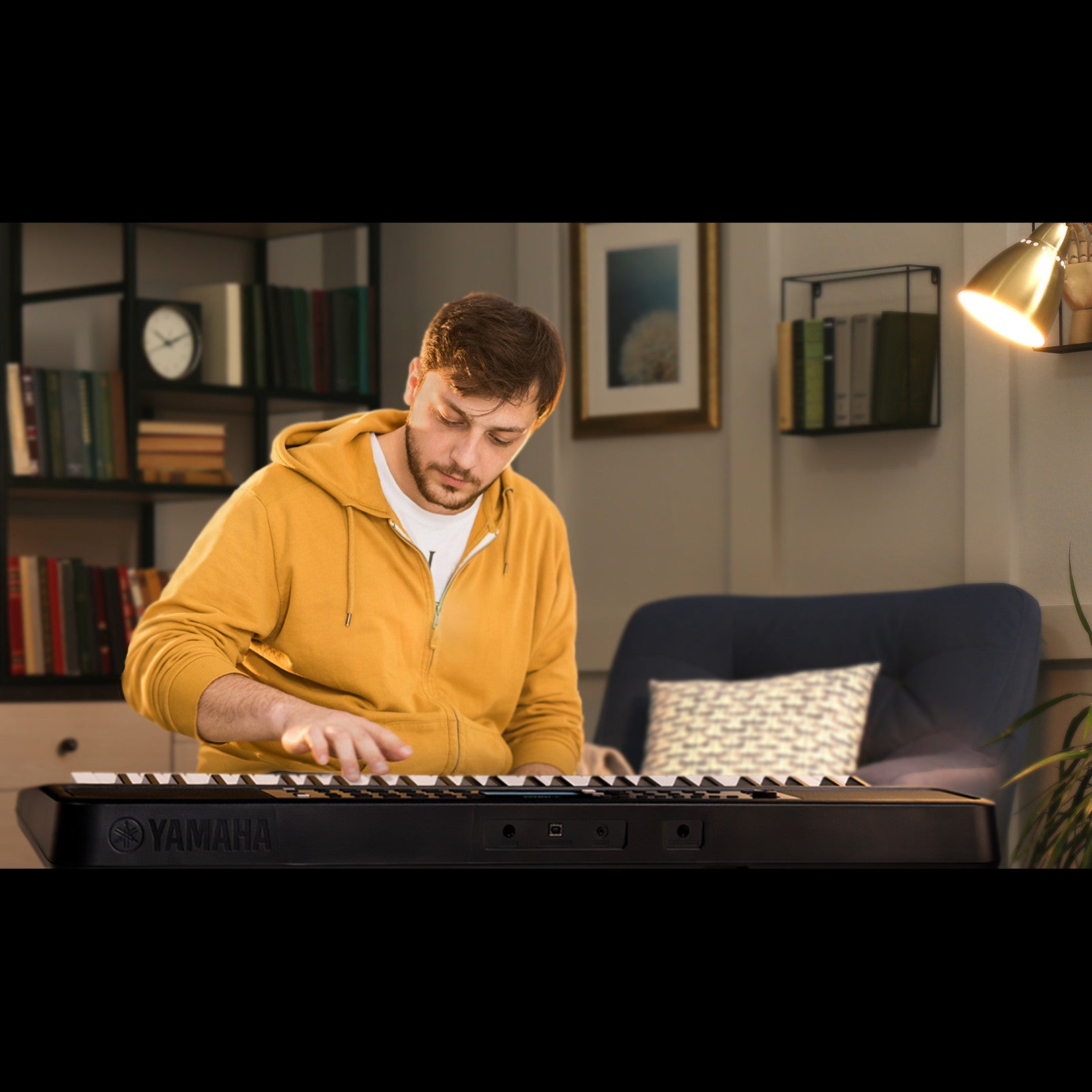Mężczyzna w żółtej bluzie z kapturem gra na czarnej klawiaturze Yamaha w przytulnym pokoju.