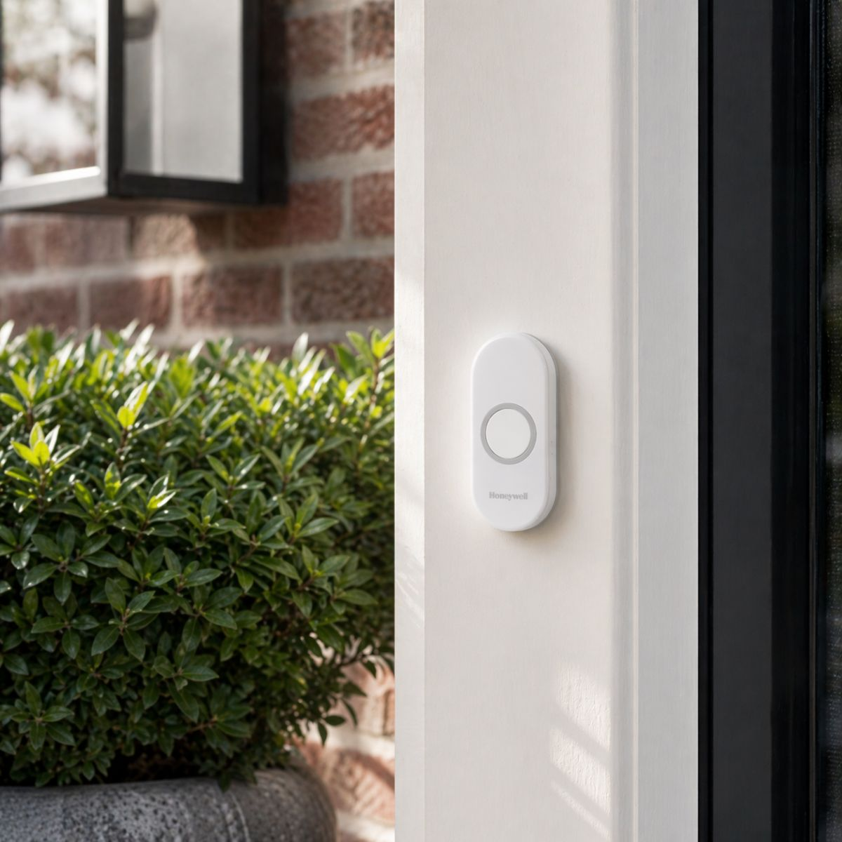 A white Honeywell doorbell button on a white door next to green bushes.
