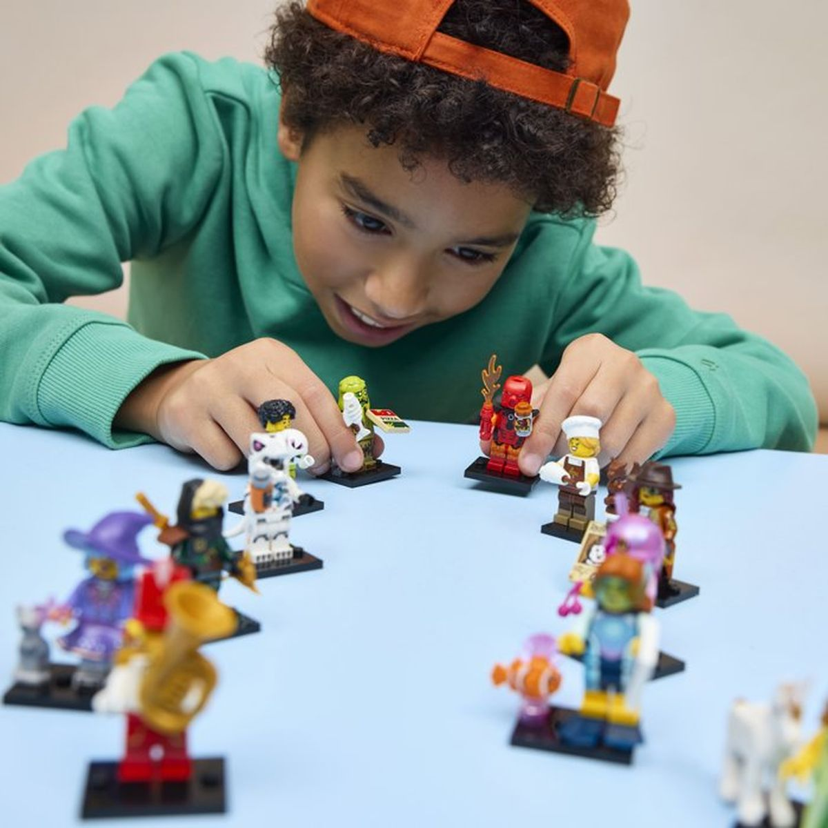 Ragazzo con cappellino arancione gioca con statuine Lego su tavolo blu.