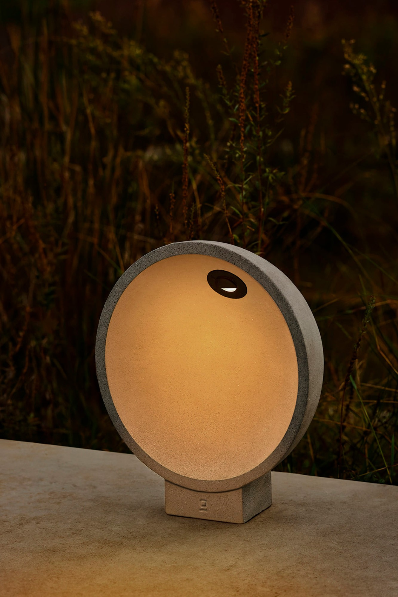 Lampada rotonda in cemento brilla calda su base di pietra, circondata da erba scura.