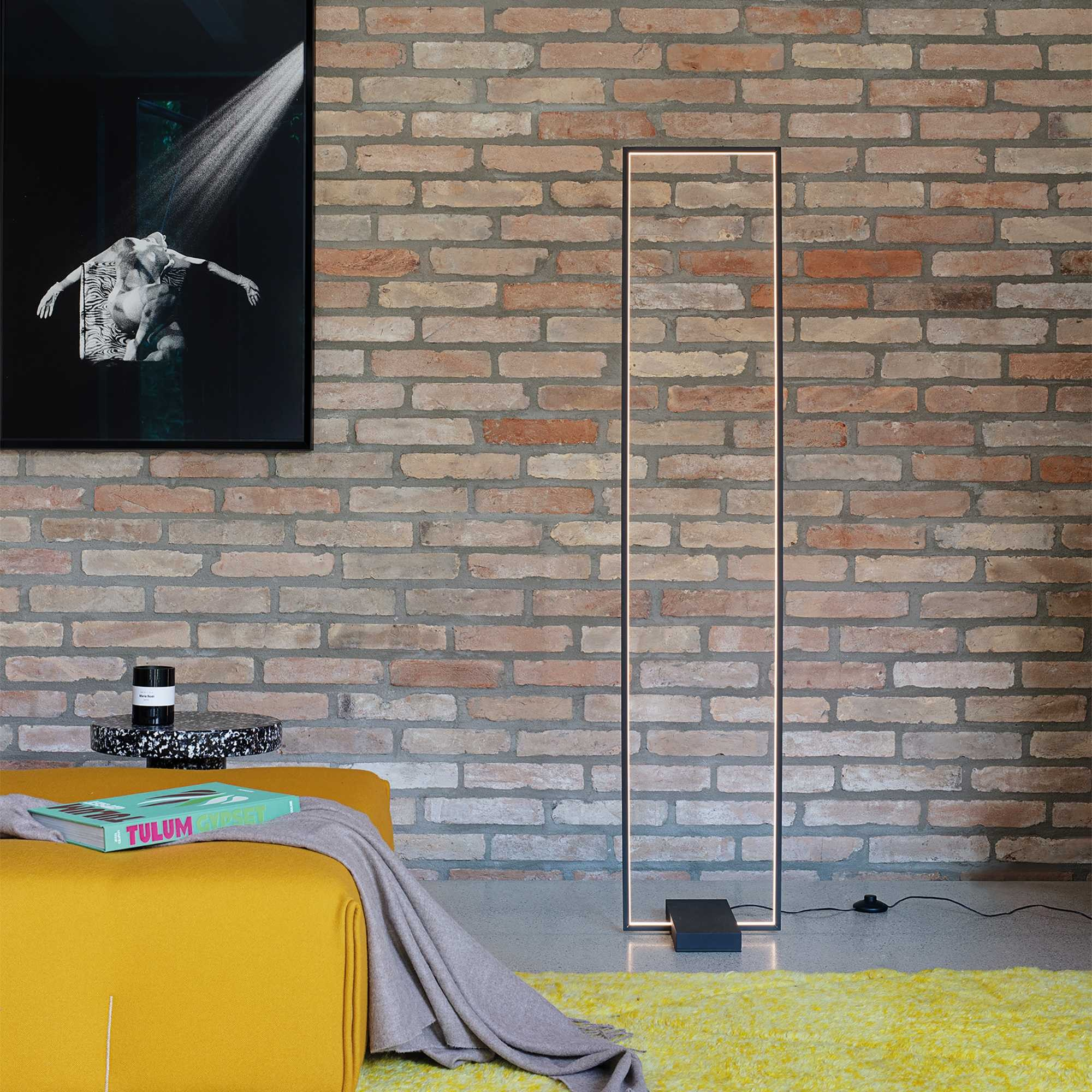 Un pouf giallo con libro e coperta, accanto a una cornice di lampada da terra rettangolare.
