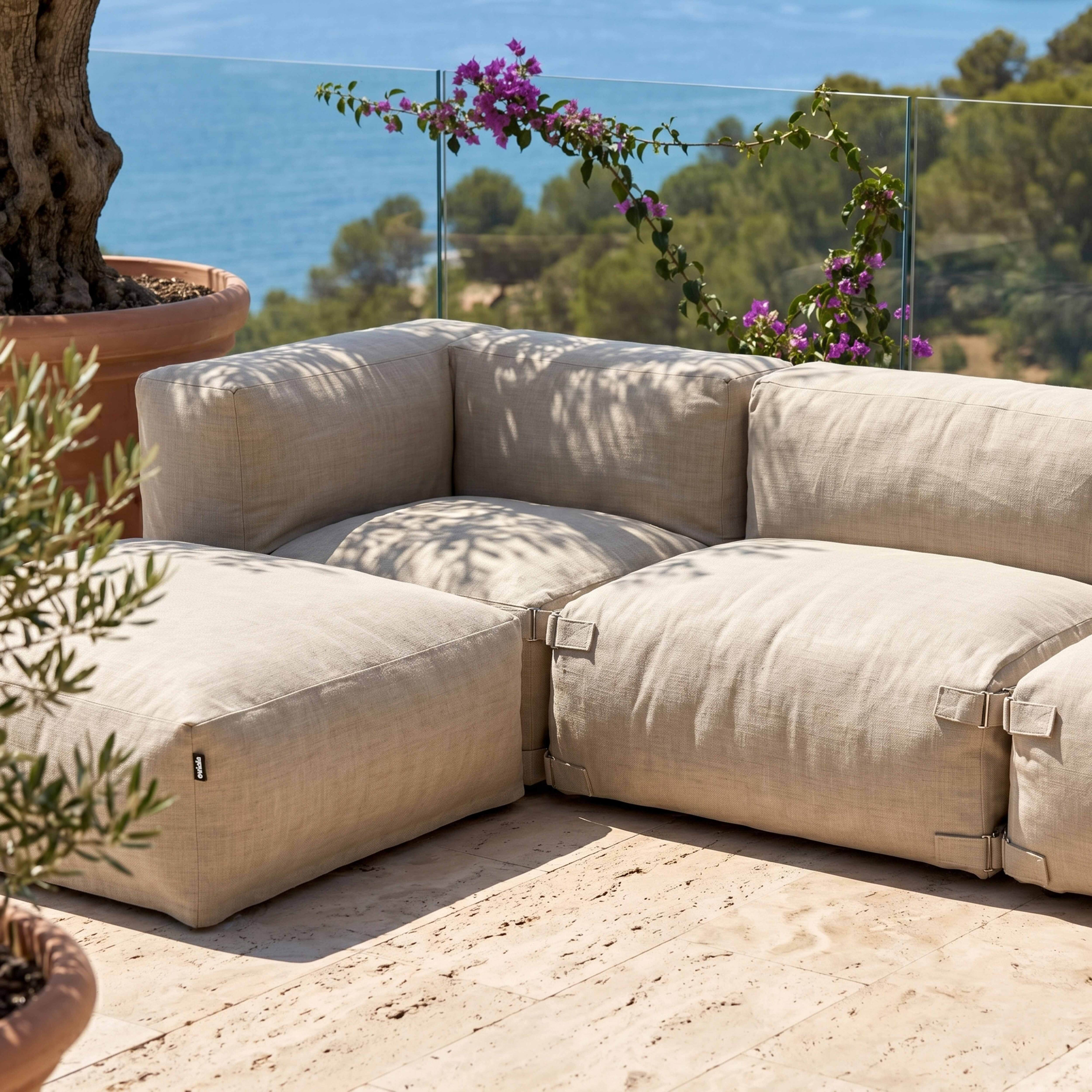 Ein beiges Ecksofa auf einer Steinterrasse mit Meerblick und Blumen.