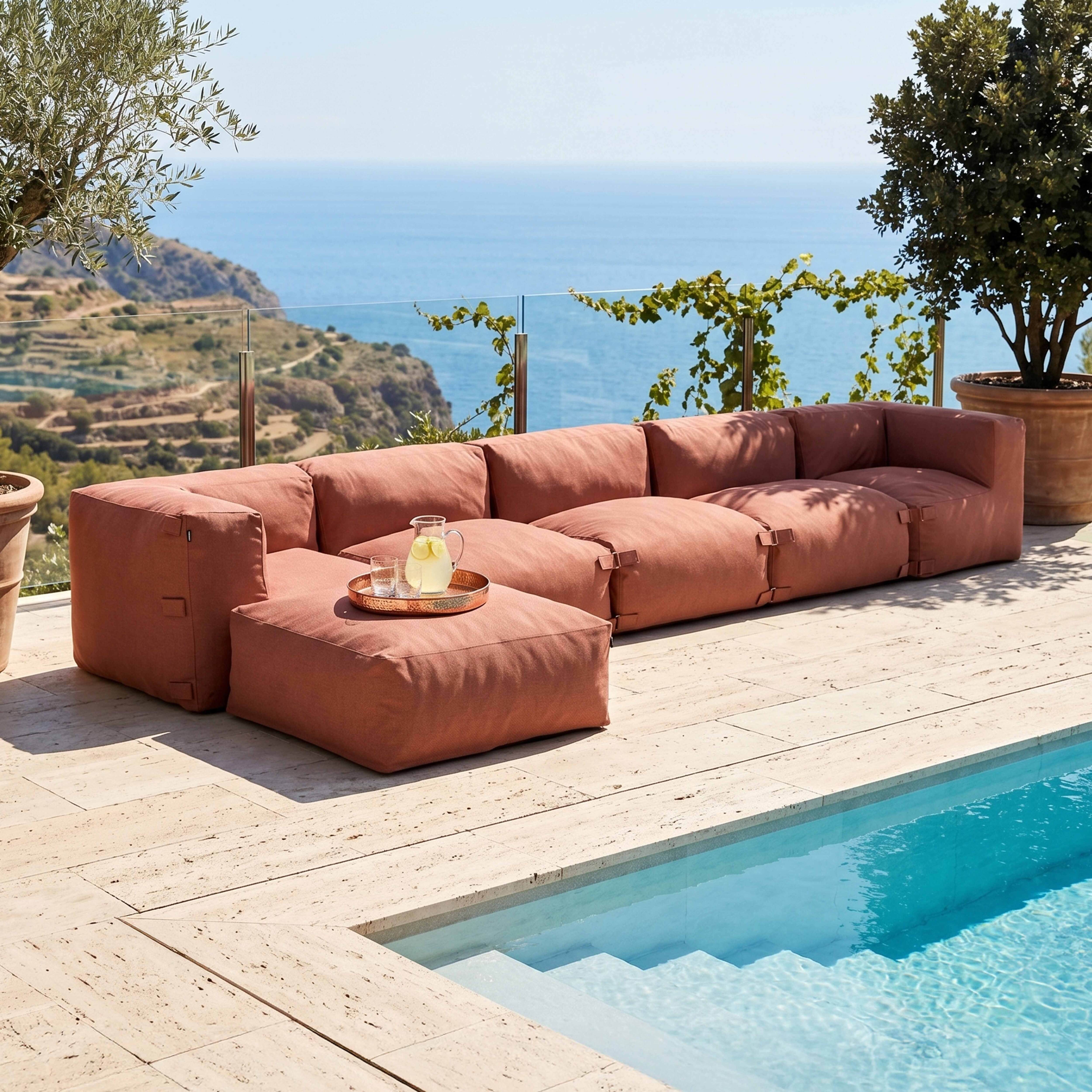 Ein korallenfarbenes Ecksofa steht auf einer Travertinterrasse neben einem Pool, mit Meer und Hügeln im Hintergrund.