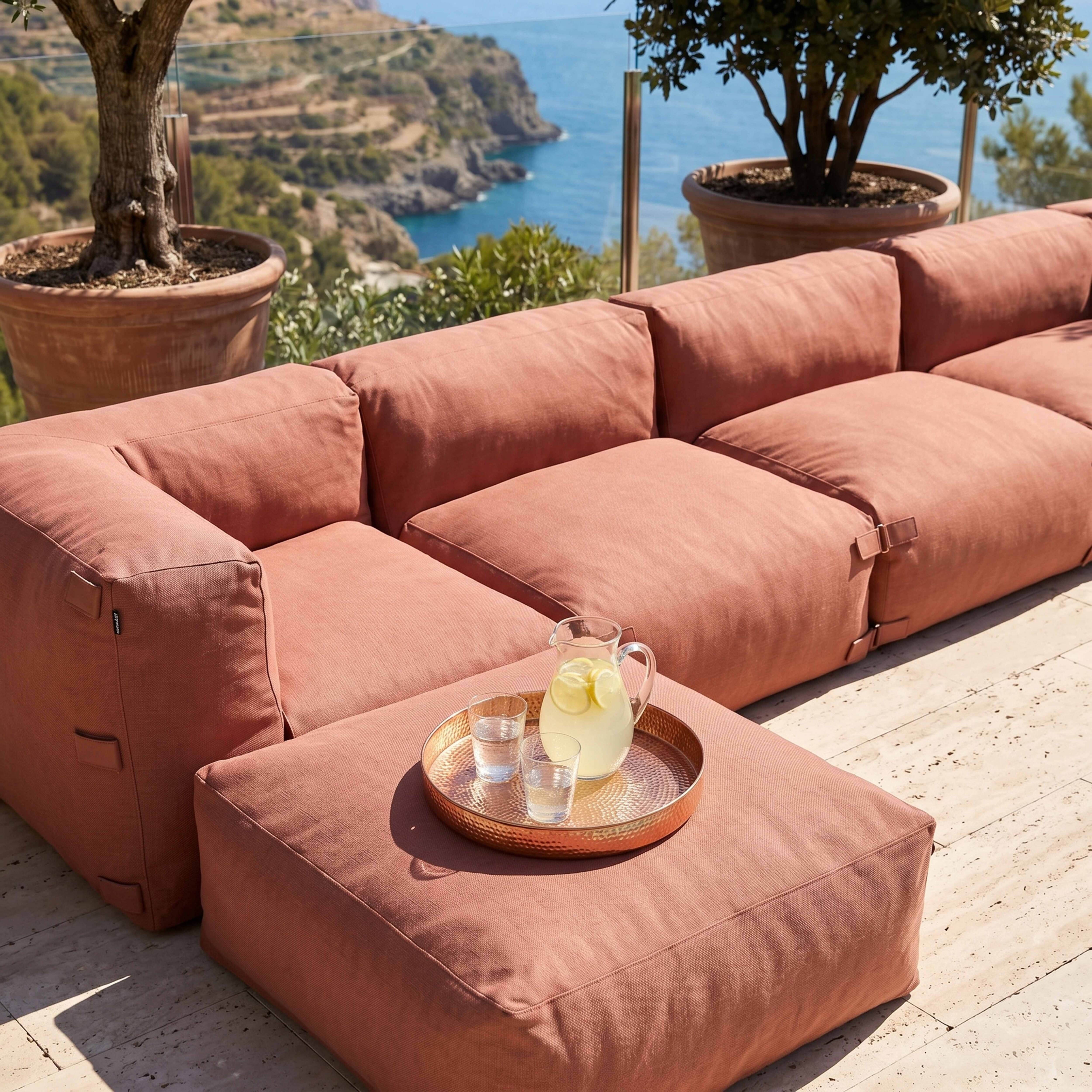 Ein korallenfarbenes Sofa mit einem Beistelltisch und einem Krug Limonade auf einer Terrasse mit Meerblick.