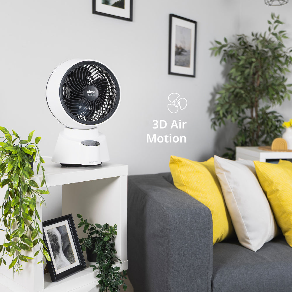 Un ventilador blanco en una estantería junto a un sofá gris con cojines amarillos.