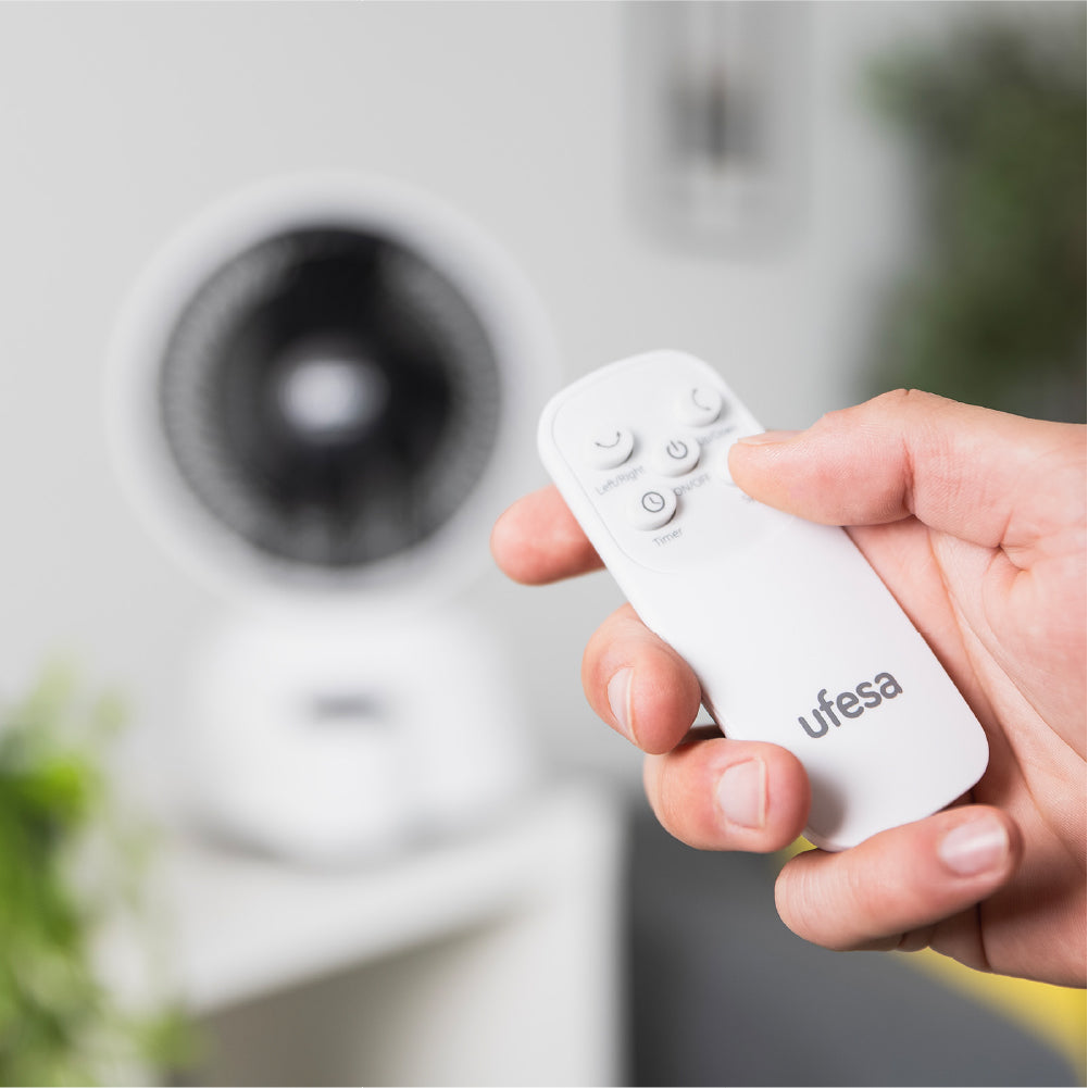 Una mano sujeta un mando a distancia blanco con botones. Un ventilador desenfocado al fondo.
