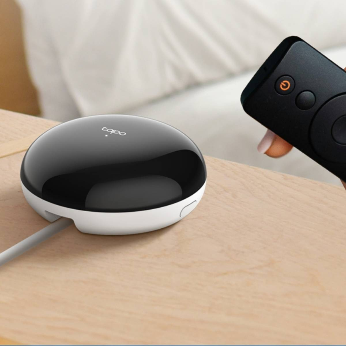 Un hub smart bianco e nero su tavolo di legno, con telecomando nero in mano.