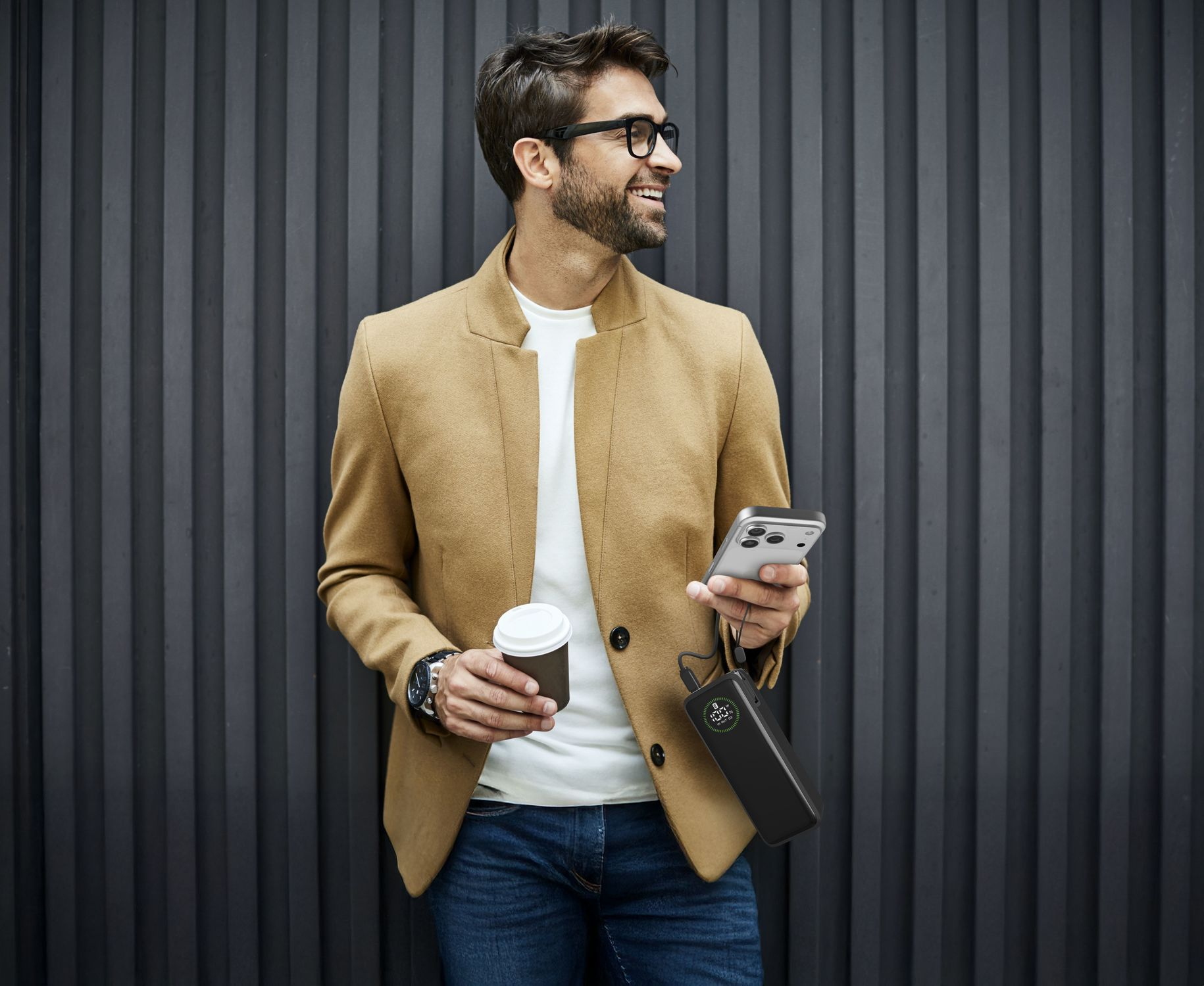 Mann mit Brille hält Kaffee und Handy vor dunklem Hintergrund.