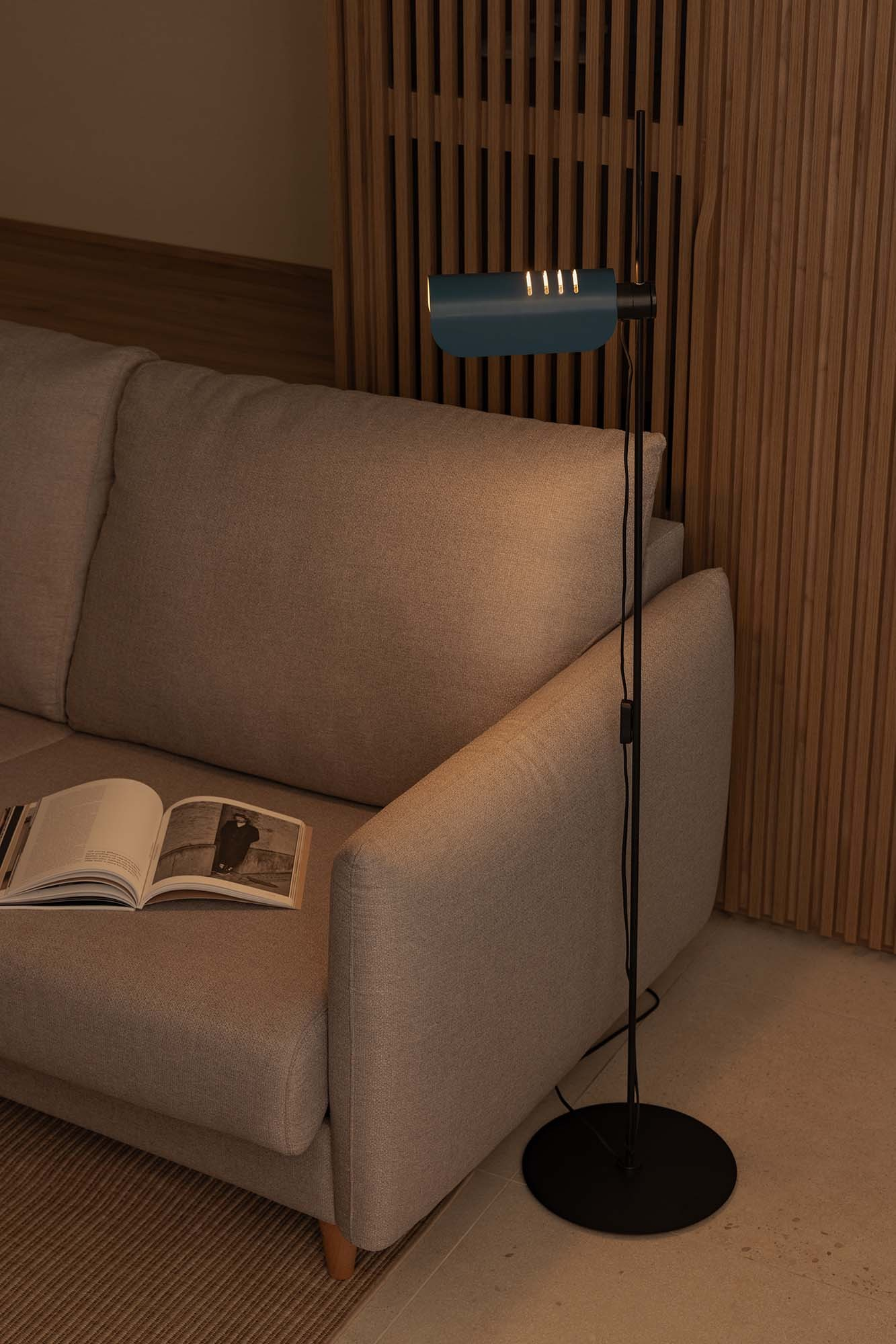 Una lampada da terra blu illumina un libro aperto su un divano beige contro una parete con pannelli di legno.