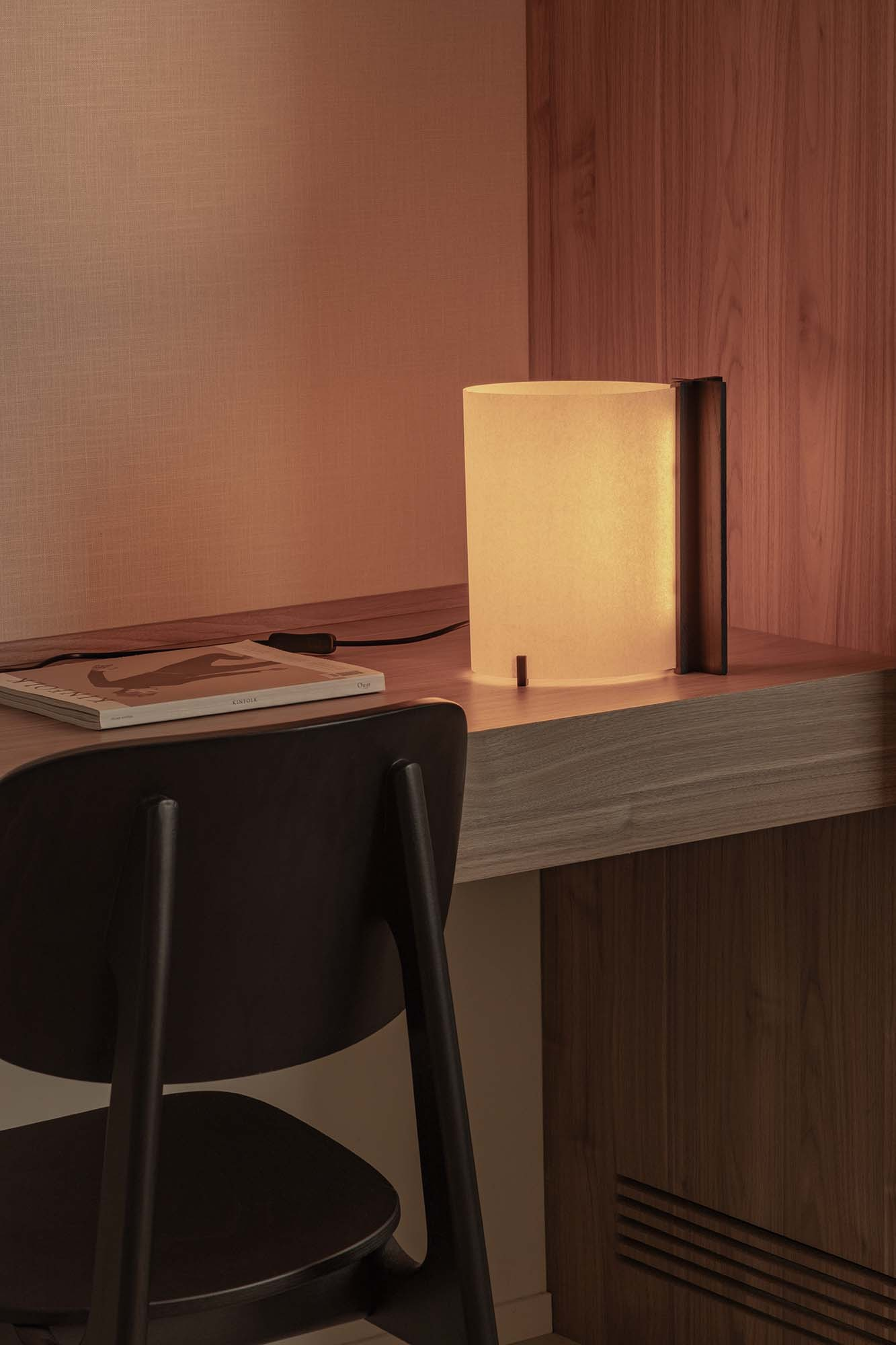 Una lampada cilindrica accesa è su una scrivania di legno accanto a una rivista. Una sedia nera in primo piano.