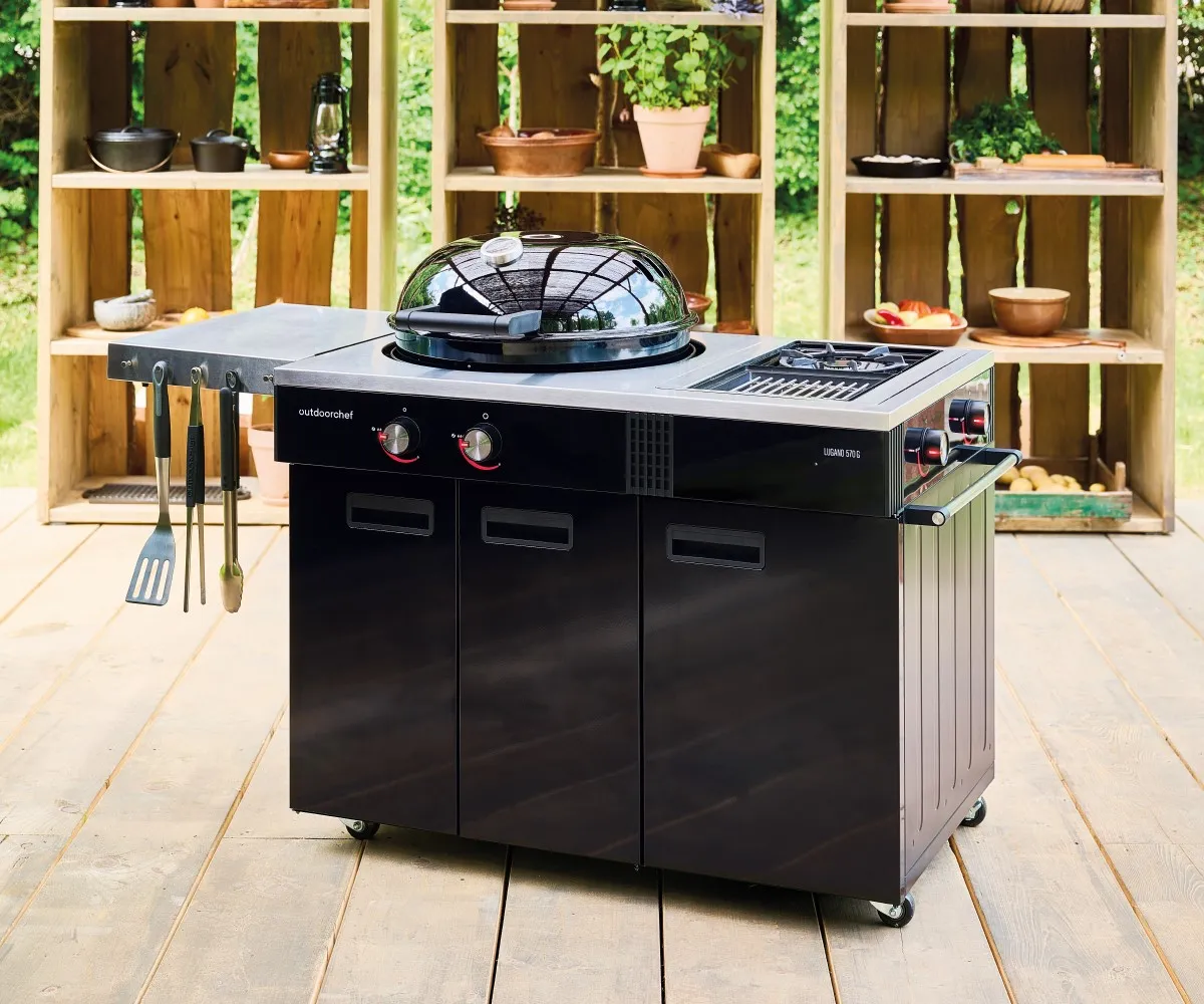 Mutfak tezgahlı siyah bir Outdoorchef ızgara ahşap güvertede duruyor, yanında raflar var.