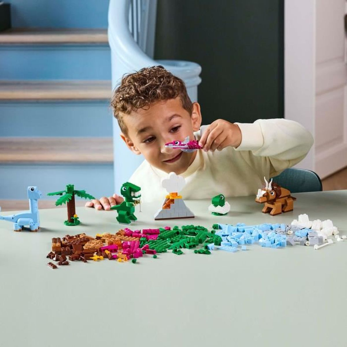 Chłopiec bawi się klockami Lego z dinozaurami i wulkanem na stole.