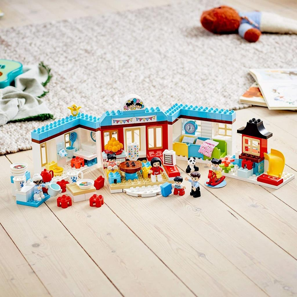 Renkli bir Lego Duplo seti, figürler ve oyuncaklarla bir binayı tasvir ediyor.