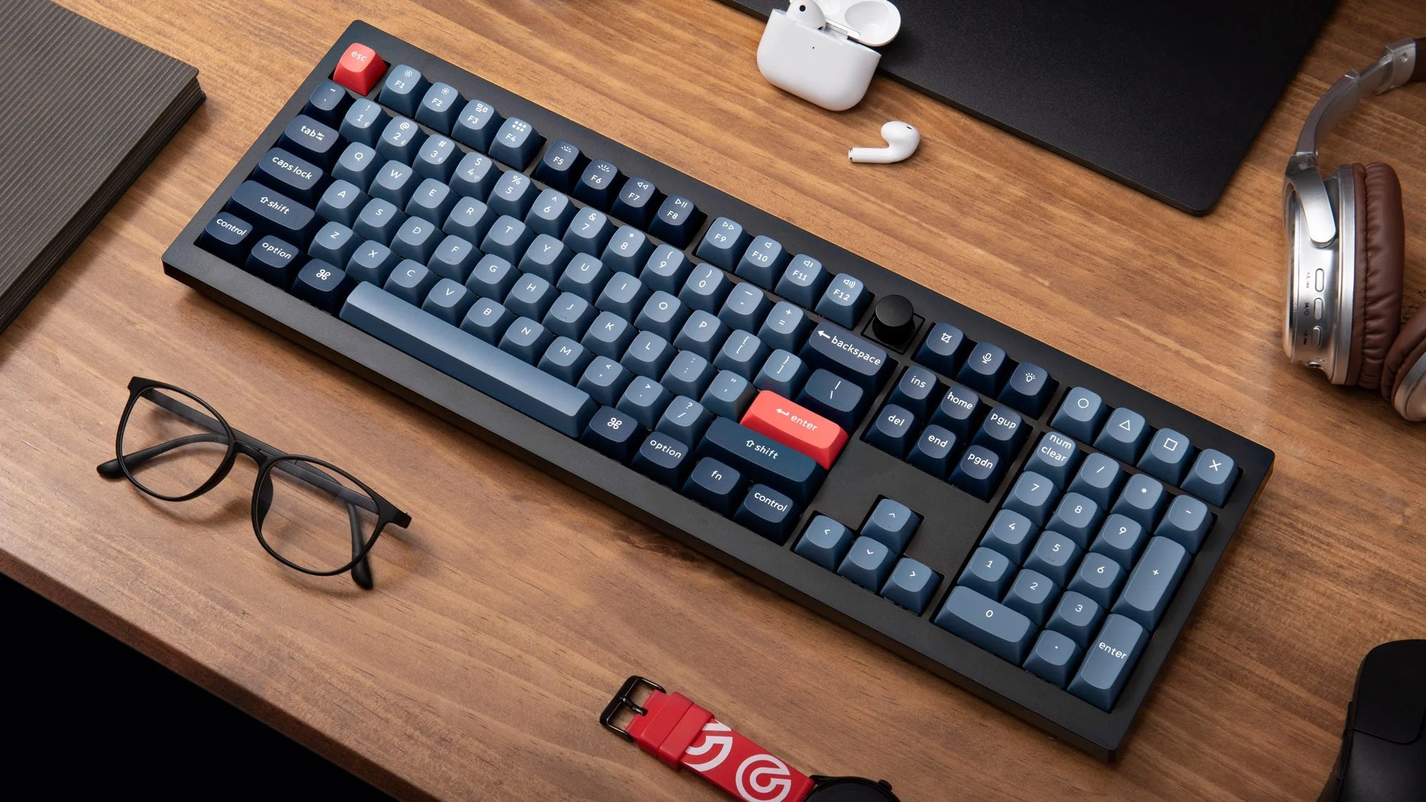 Eine schwarze mechanische Tastatur mit blauen Tasten, Brille, Kopfhörer und Ohrhörer auf einem Holztisch.