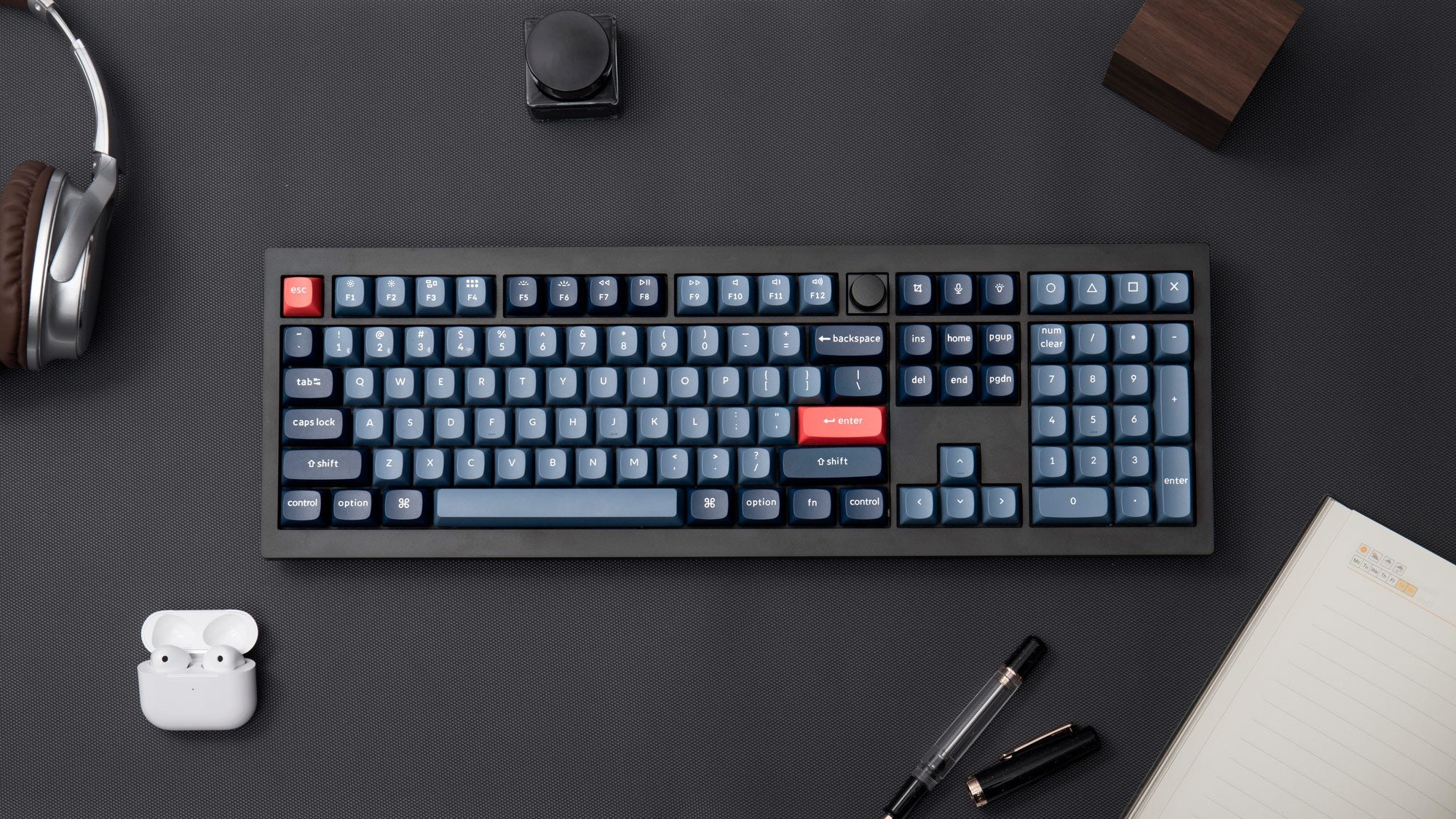 Eine Tastatur, Kopfhörer, AirPods, ein Notizbuch und ein Stift auf einer Matte.