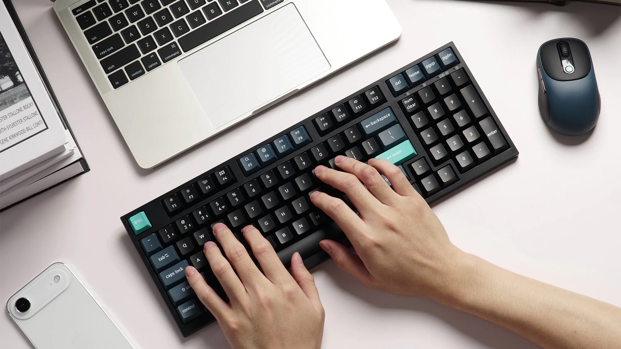 Ręce piszące na klawiaturze obok laptopa, myszy i smartfona.