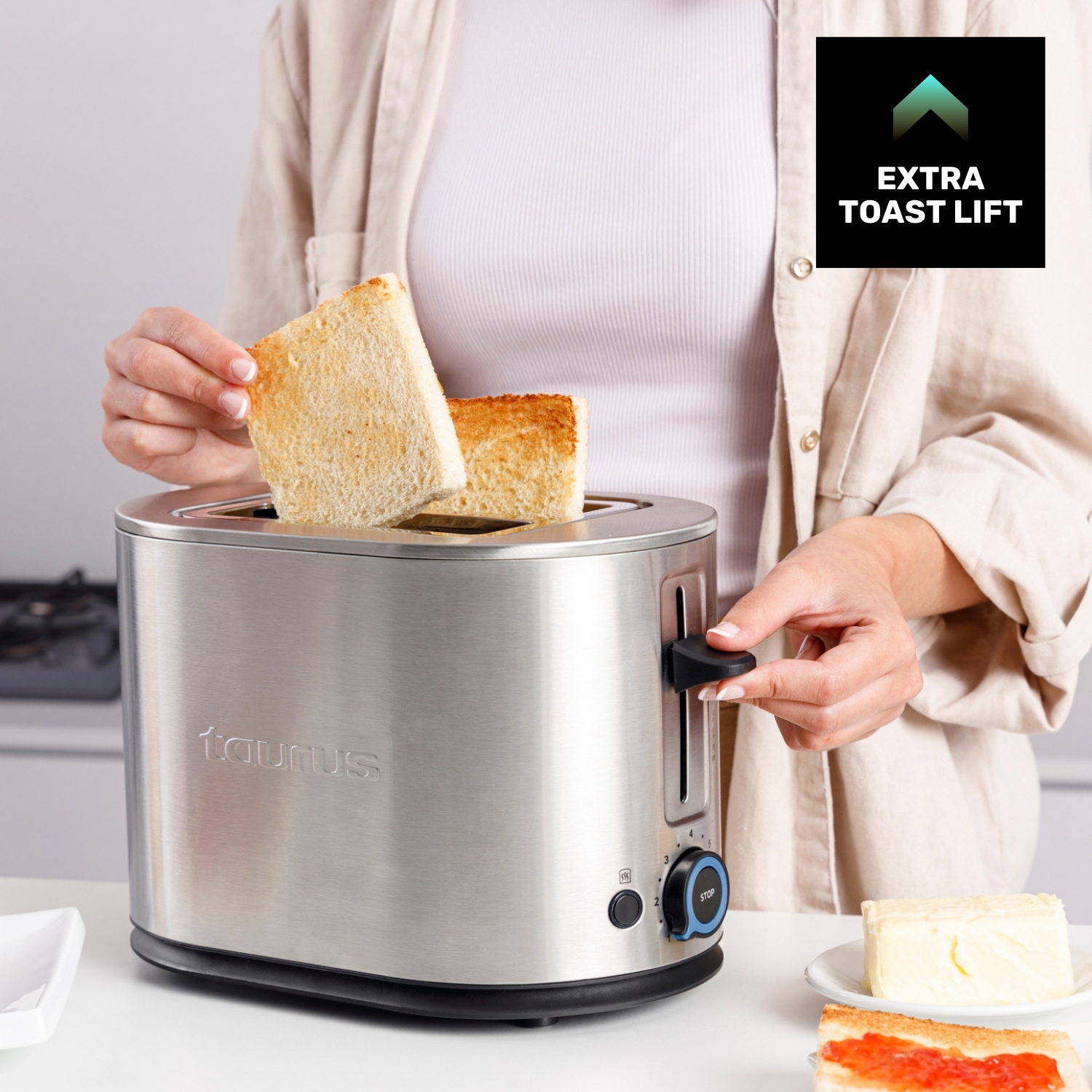 Una persona saca tostadas de una tostadora plateada. Cerca hay mantequilla y tostadas con mermelada.