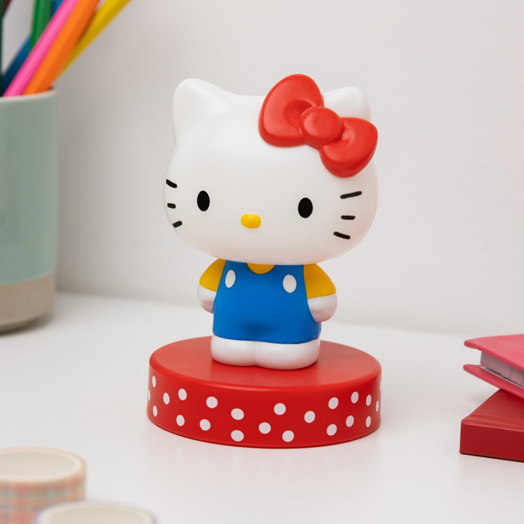 Figurka Hello Kitty z czerwoną kokardką, niebieskimi ogrodniczkami i żółtą bluzką.