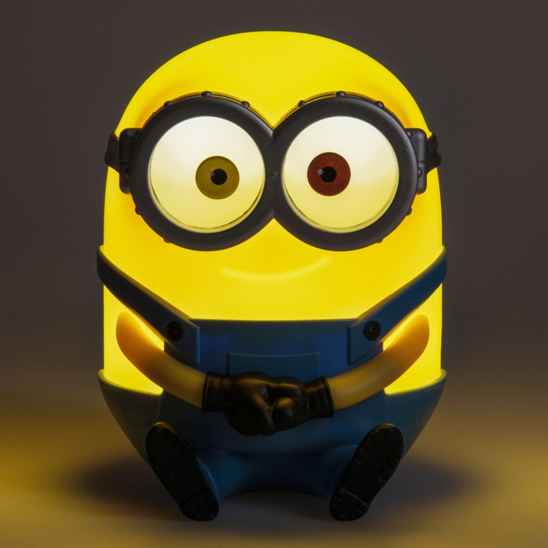 Żółta lampa Minion w niebieskich ogrodniczkach siedzi ze złożonymi dłońmi.