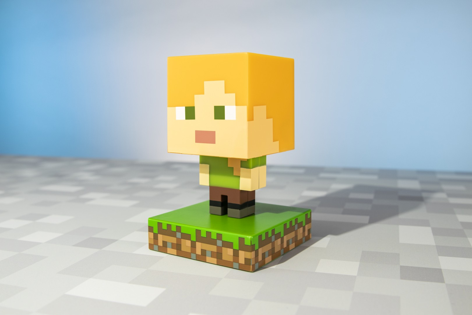 Pikselowa figurka Alexa z Minecrafta z pomarańczowymi włosami, zieloną koszulką i brązowymi spodniami na podstawie bloku trawy.
