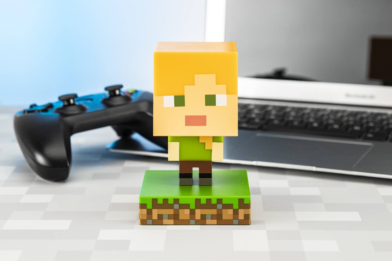 Figurka Minecraft Alex na zielonej podstawie z trawą, obok kontroler i laptop.