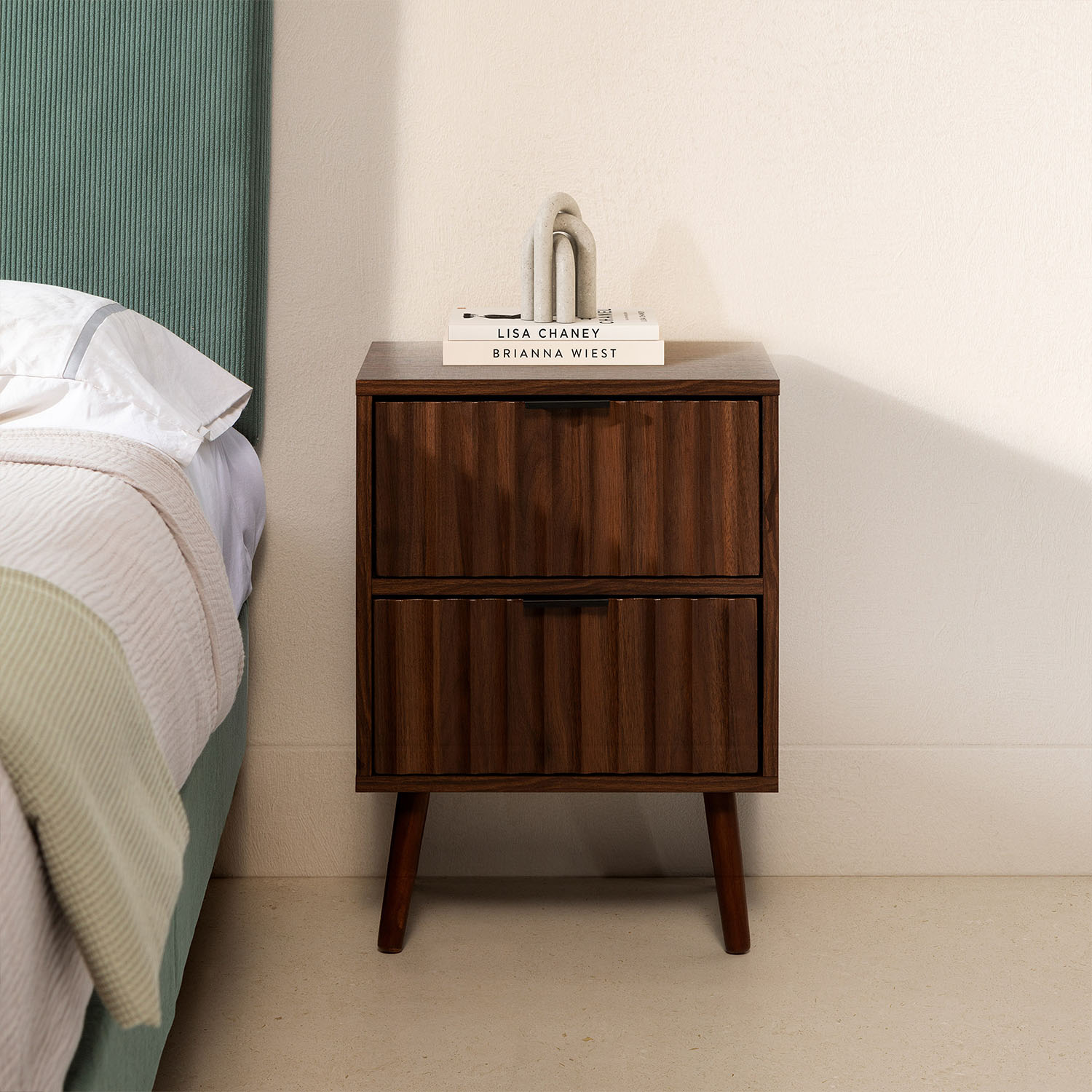 Una mesita de noche de madera con dos cajones se encuentra junto a una cama con ropa de cama blanca.