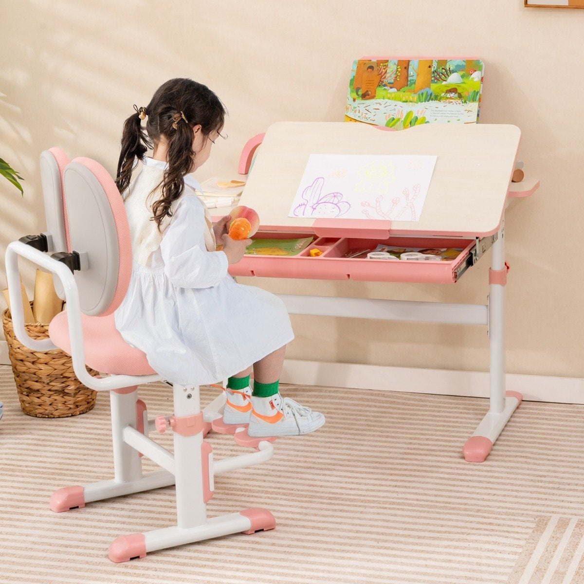 Una niña se sienta en un escritorio rosa ajustable, dibujando con crayones. Un libro está abierto.