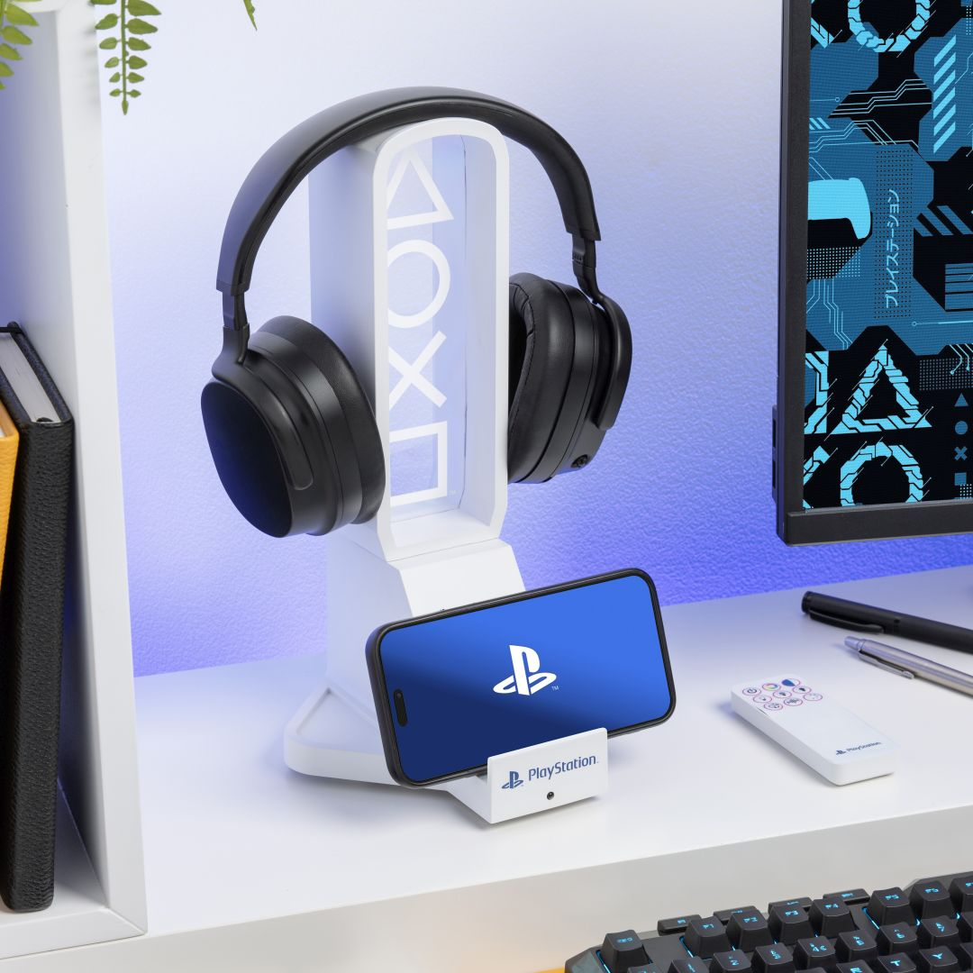 Schwarze Kopfhörer auf weißem Ständer mit PlayStation-Logo, daneben ein Handy und eine Fernbedienung.