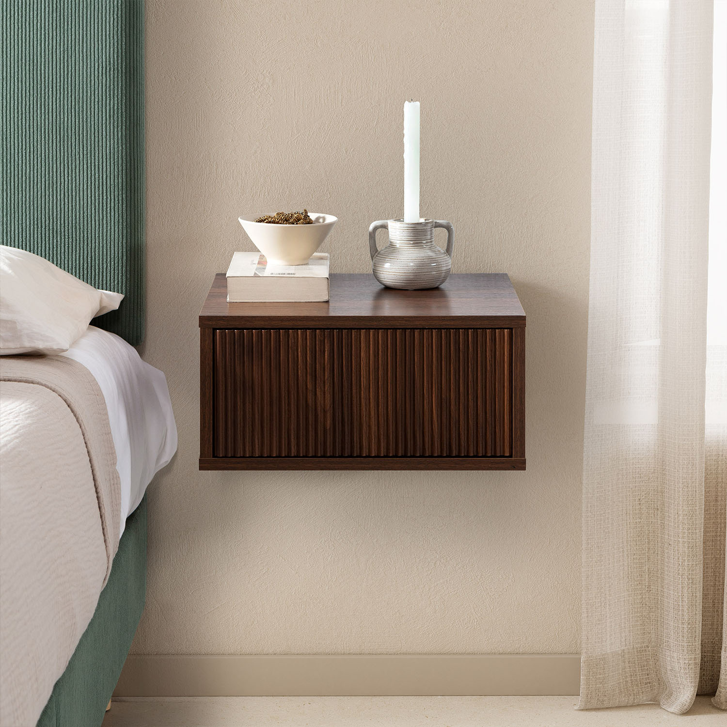 Una mesita de noche de madera oscura con frente acanalado junto a una cama con cabecero verde.