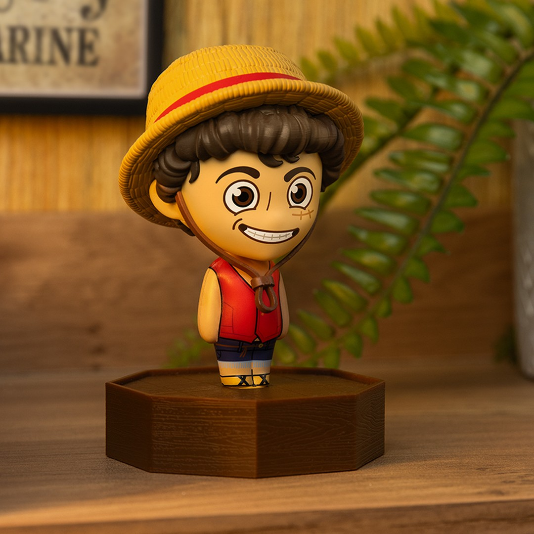 Figurka chibi chłopca w słomkowym kapeluszu i czerwonej kamizelce stoi na brązowej podstawie.