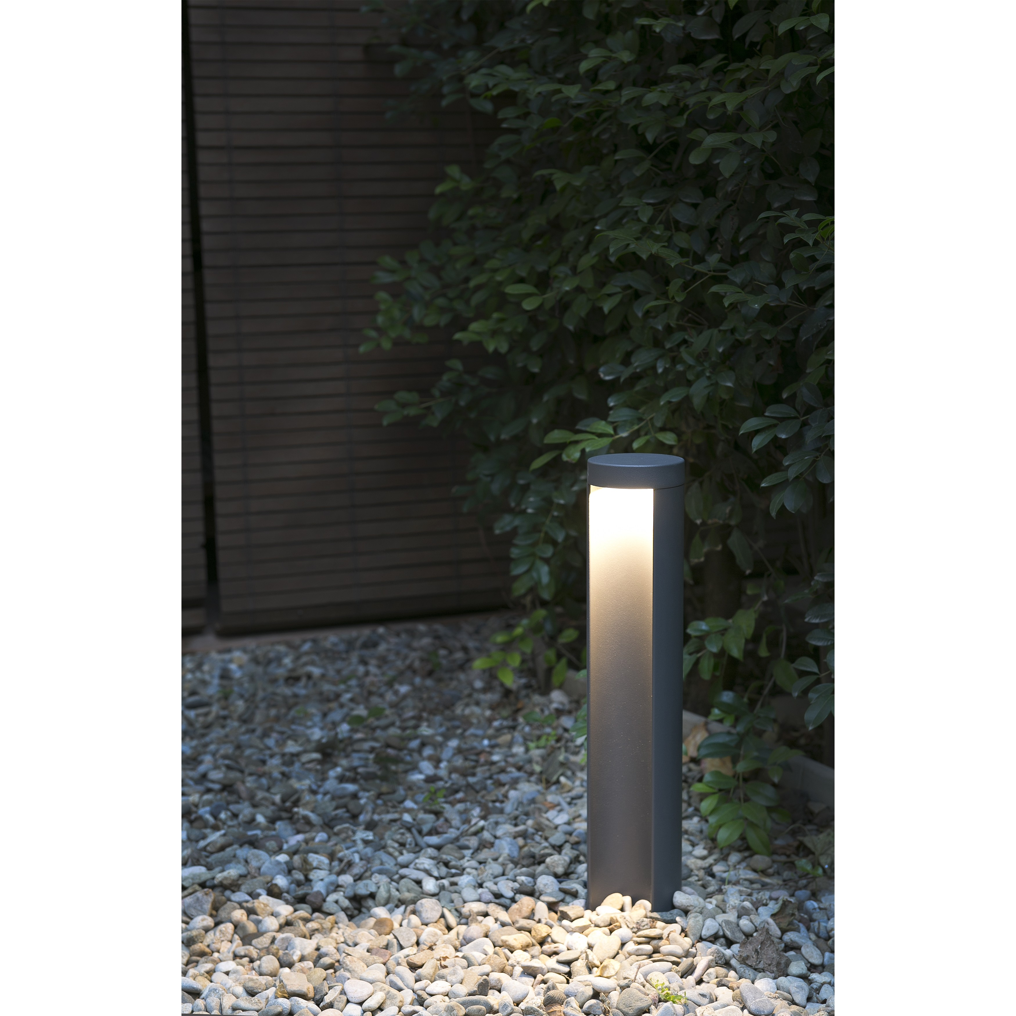 Un lampione moderno grigio illumina un sentiero di ciottoli e lussureggianti cespugli verdi.