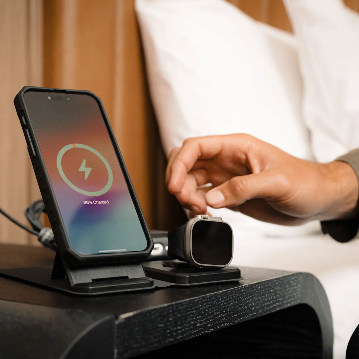 Dłoń kładzie Apple Watch na ładowarce obok telefonu na stojaku.