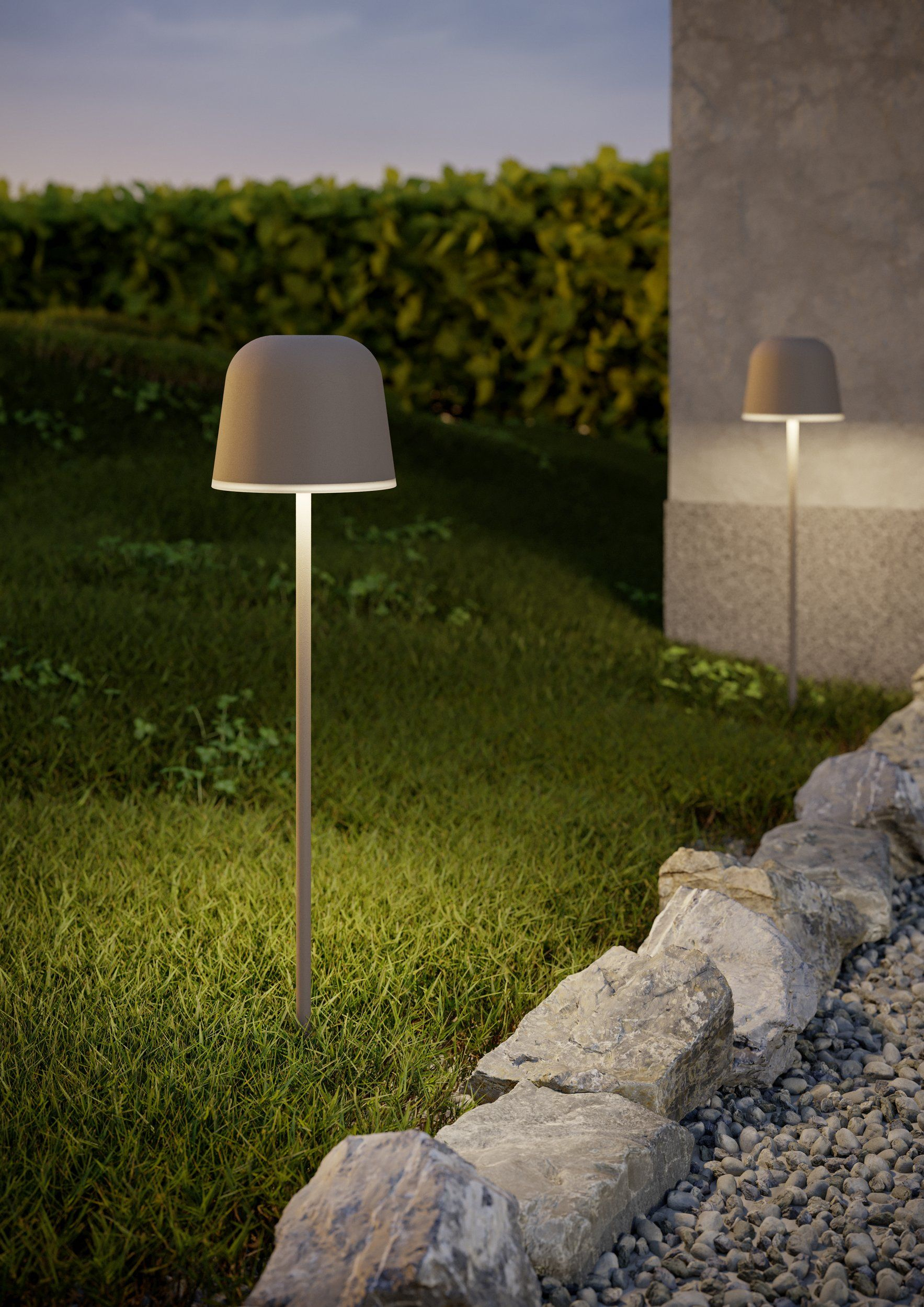 Zwei moderne Außenlampen beleuchten einen grasbewachsenen Hof mit Steinen und einer Hecke.