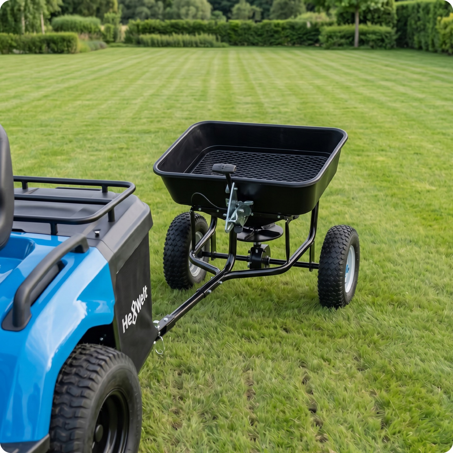 Ein blauer Rasentraktor zieht einen schwarzen Streuwagen auf einem gepflegten Rasen.