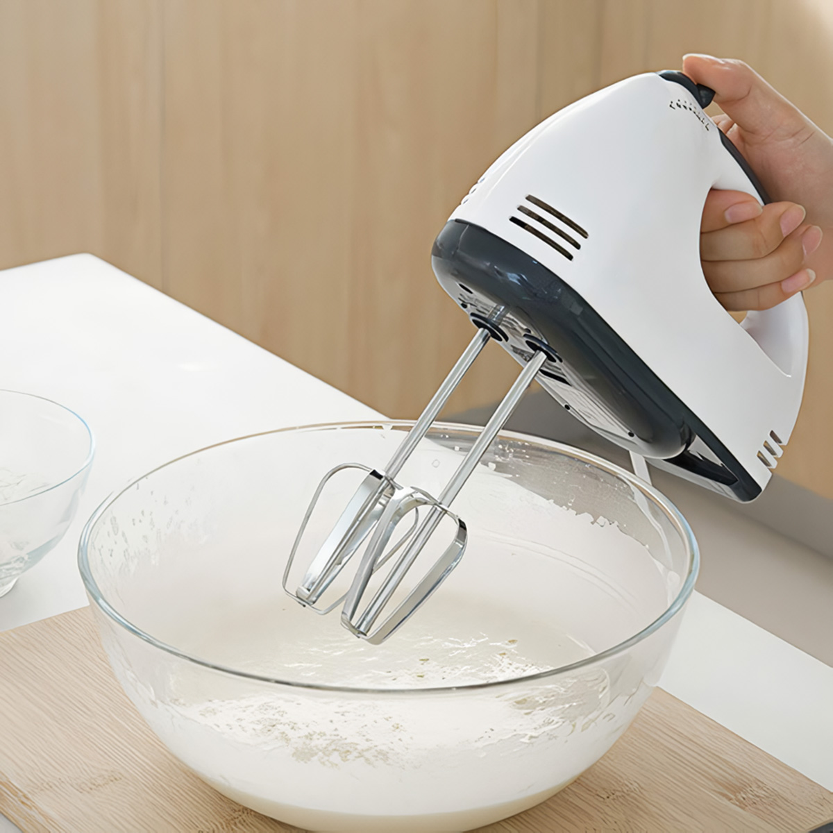 Una mano tiene uno sbattitore elettrico bianco e grigio sopra una ciotola di vetro con pastella.