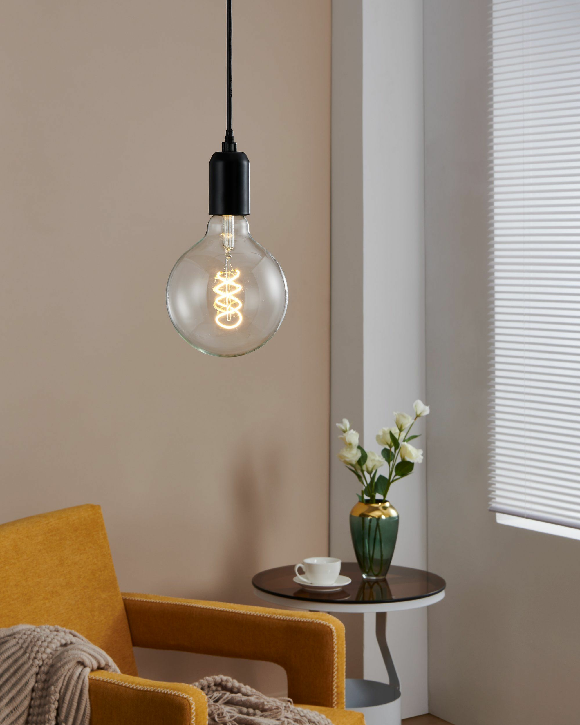 Una lampadina a sospensione sopra una poltrona gialla, con un tavolino e un vaso nelle vicinanze.