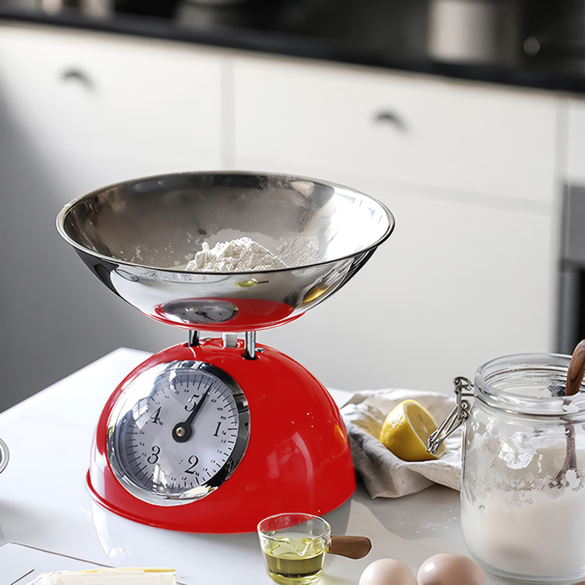 Una báscula de cocina roja con bol de acero inoxidable, con harina. Huevos, gajo de limón y harina en un frasco cerca.