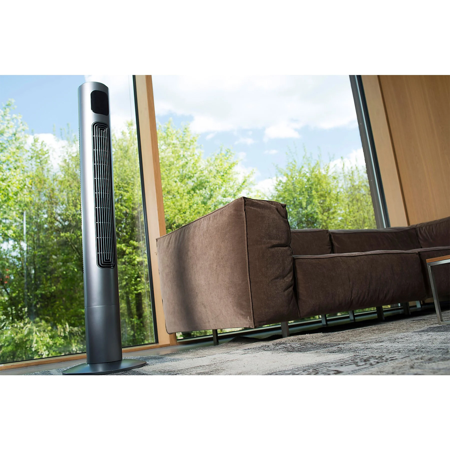 Ein schlanker, silberner Turmventilator steht neben einem braunen Ecksofa vor einem großen Fenster.