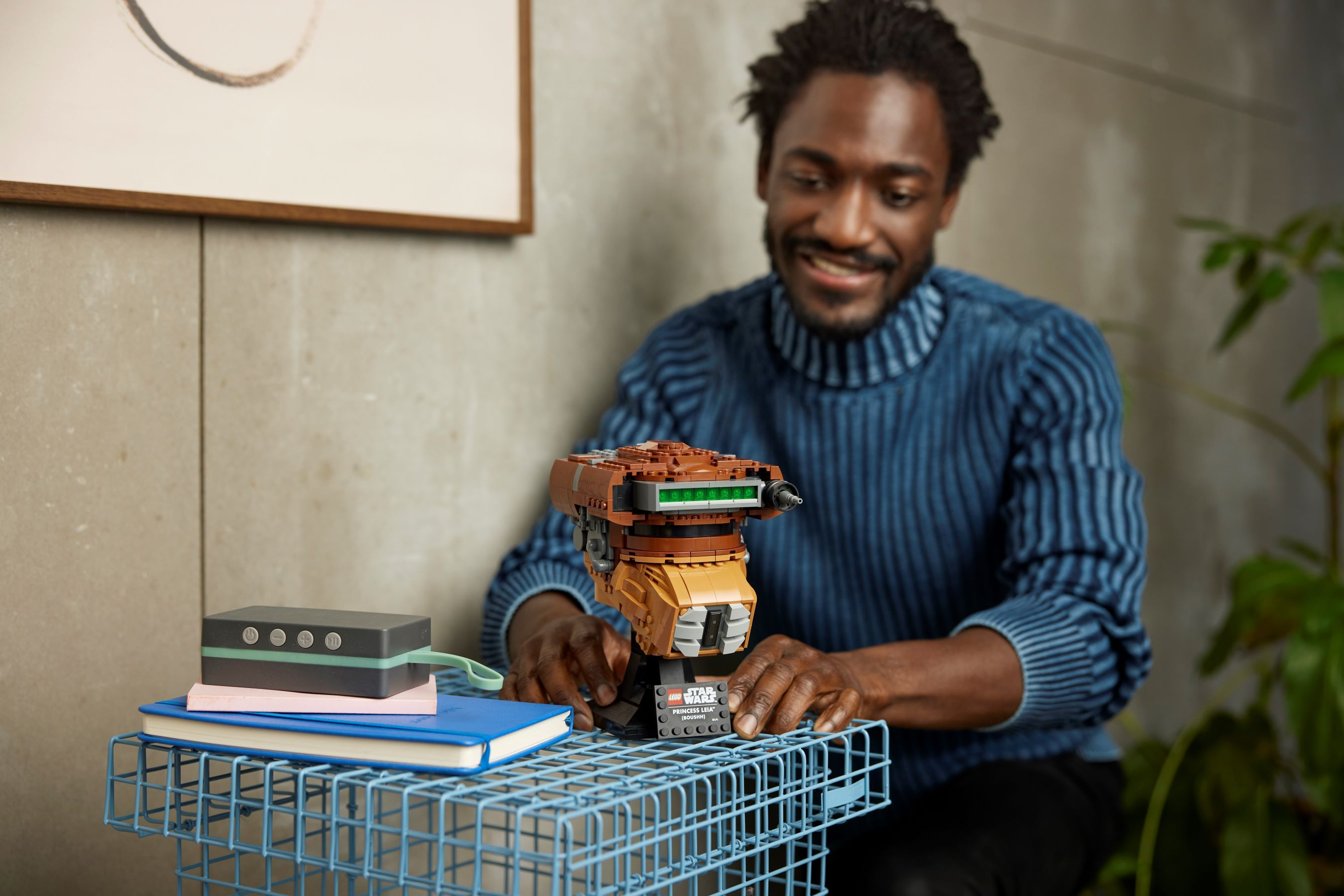 Mann im blauen Pullover baut einen Star Wars Lego Droiden. Bücher und Lautsprecher in der Nähe.