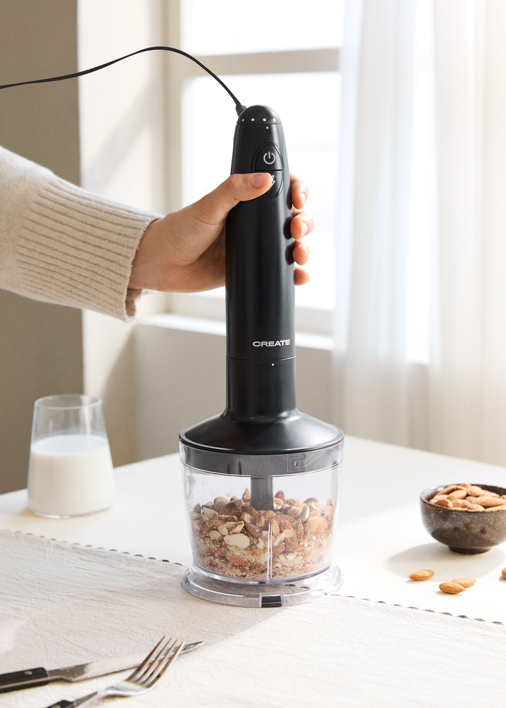 Una mano sostiene una batidora de inmersión negra sobre un recipiente con nueces picadas. Cerca hay un vaso de leche y un bol de almendras.