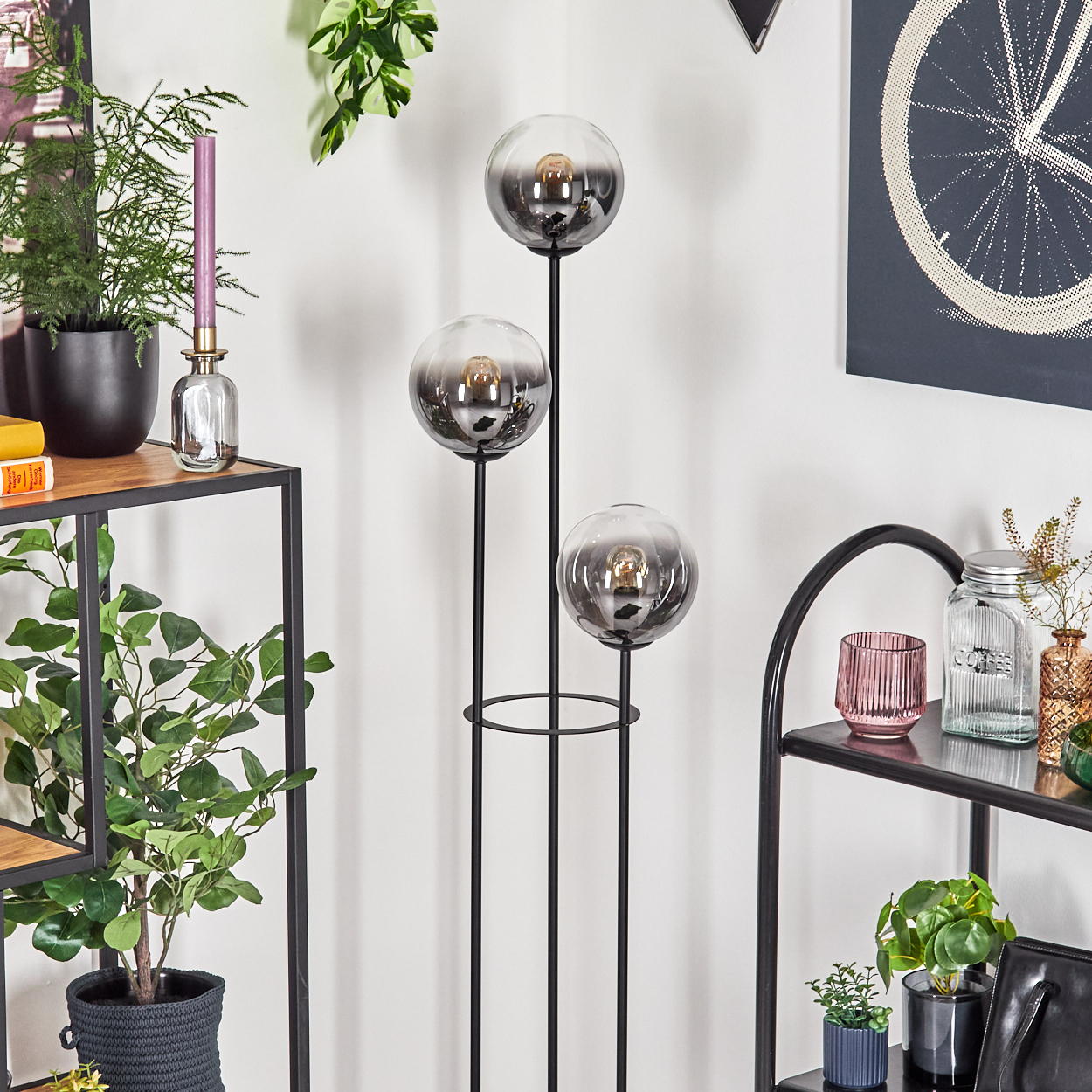 Eine moderne Stehlampe mit drei rauchglasigen Kugeln auf schwarzen Stielen. Pflanzen und Deko auf Regalen.