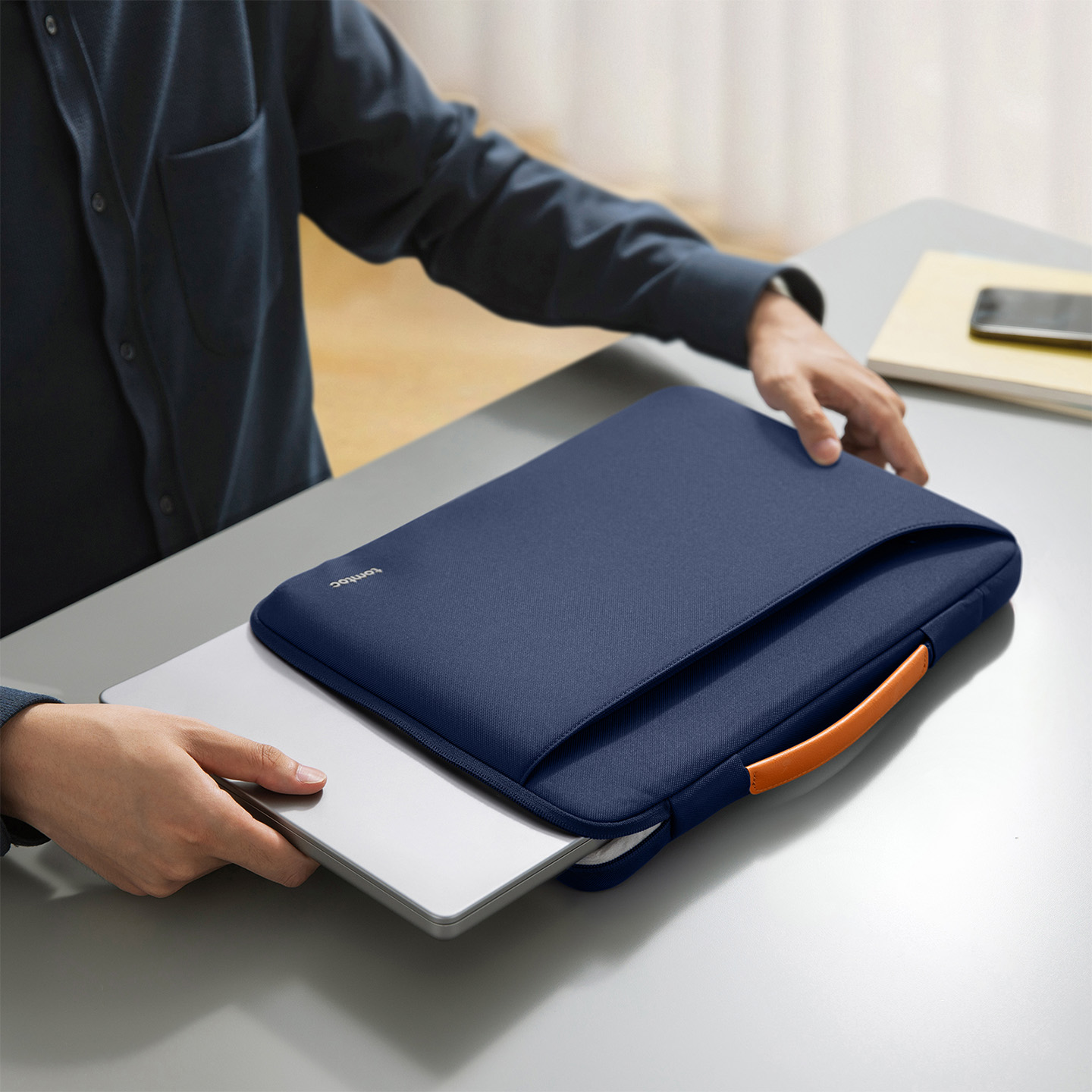 Bir kişi turuncu saplı lacivert laptop kılıfına gümüş rengi bir dizüstü bilgisayar yerleştiriyor.