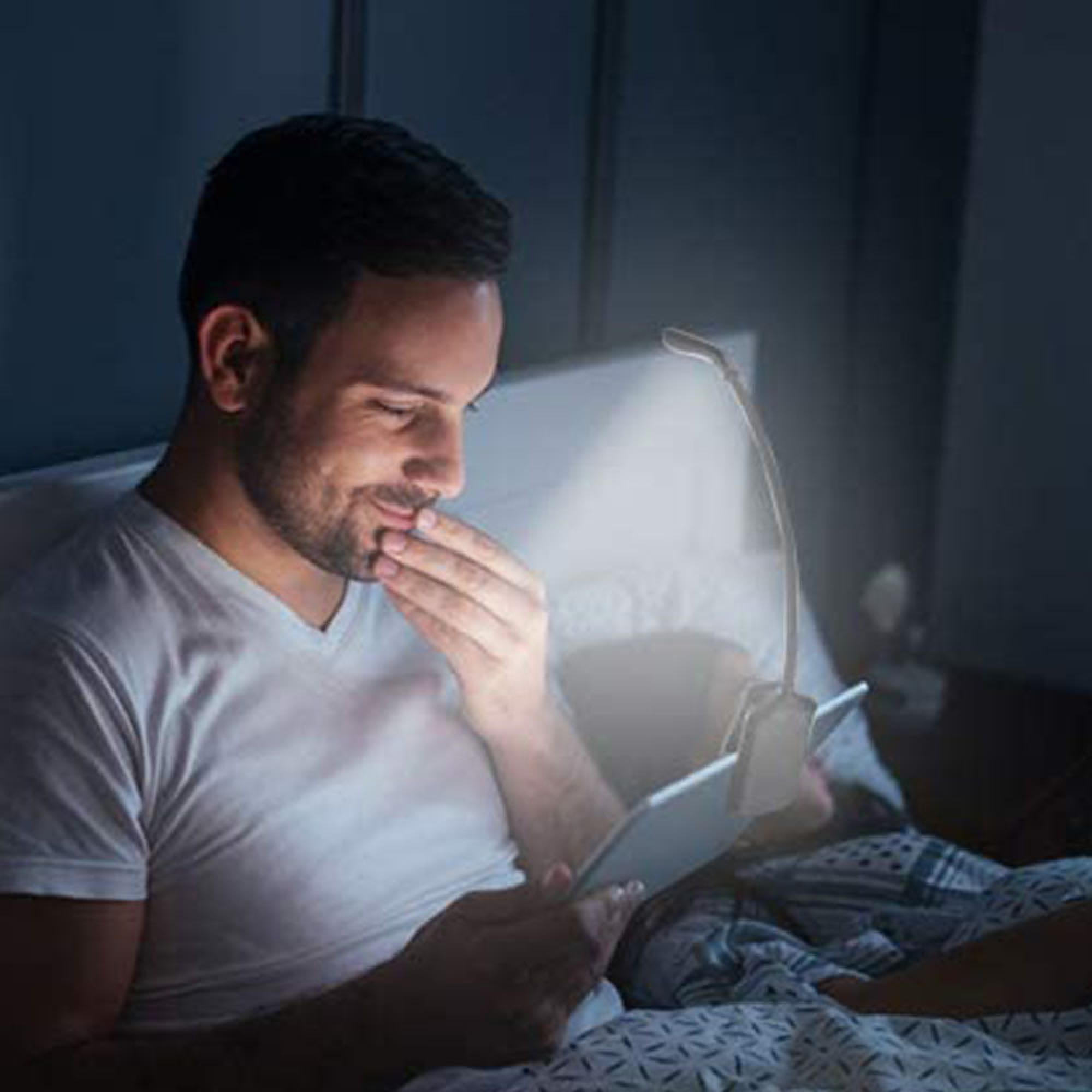Mann im Bett liest Tablet unter Klemmleuchte. Dunkler Raum.