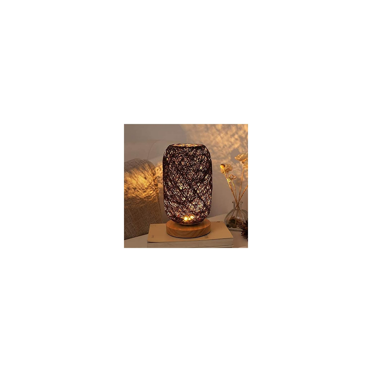 Eine gewebte Lampe mit Holzsockel leuchtet auf einem Buch neben einem Kissen und einer Vase.