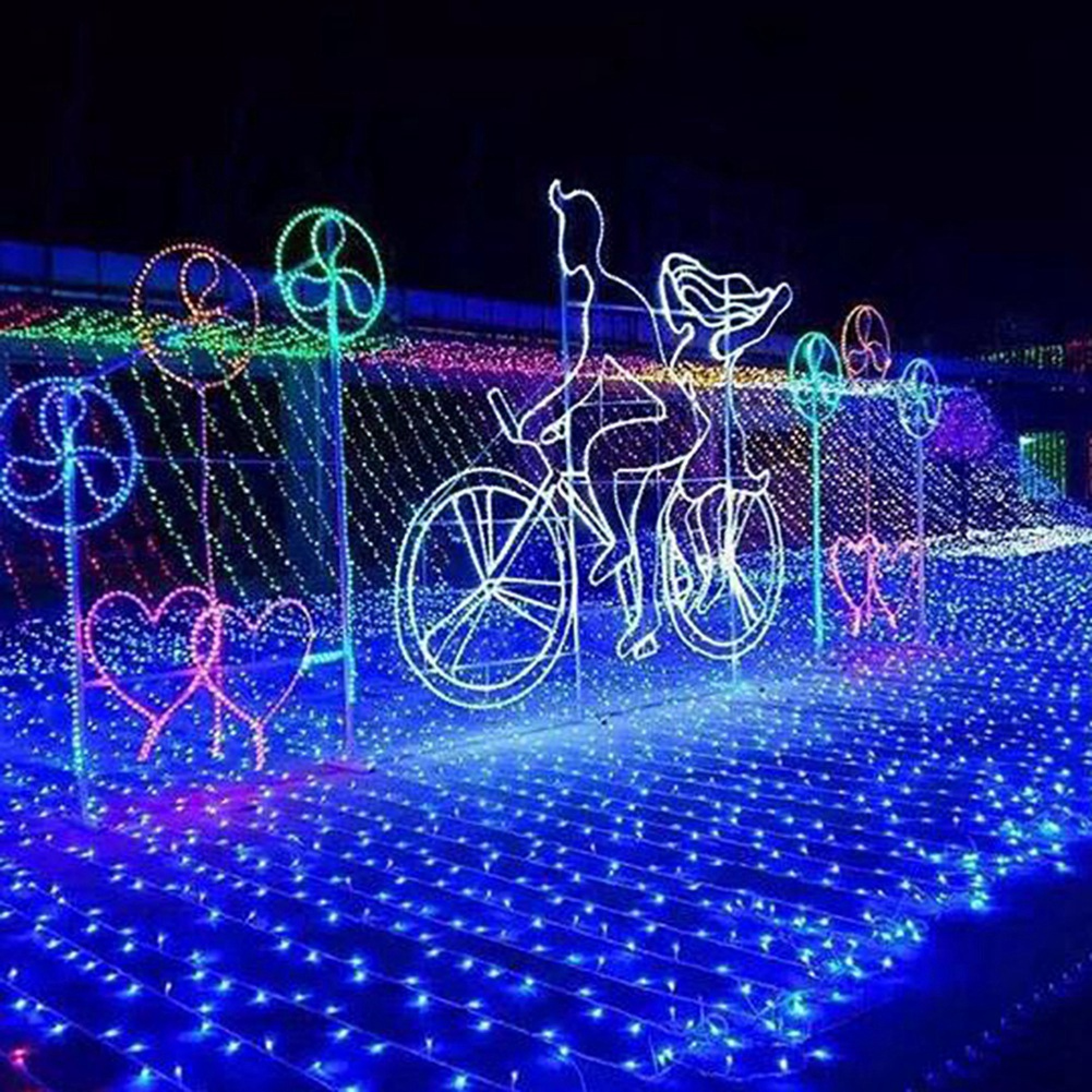 Nachtszenen mit weißen Lichtumrissen eines Paares auf einem Fahrrad, umgeben von bunten Lichtern.