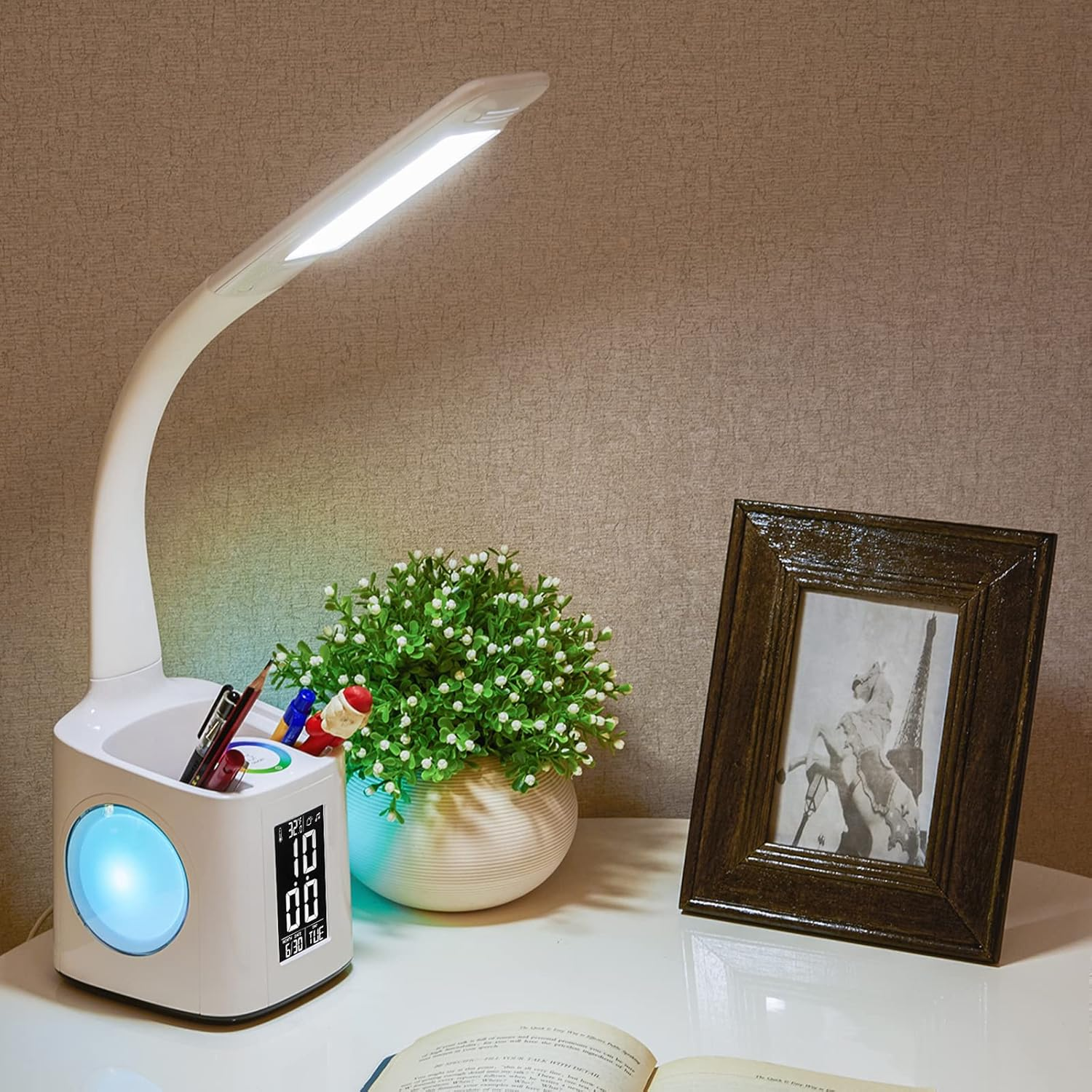 Weiße Schreibtischlampe mit Stiftehalter, Uhr, Kalender und blauem Licht. Daneben eine Topfpflanze und ein Bilderrahmen.