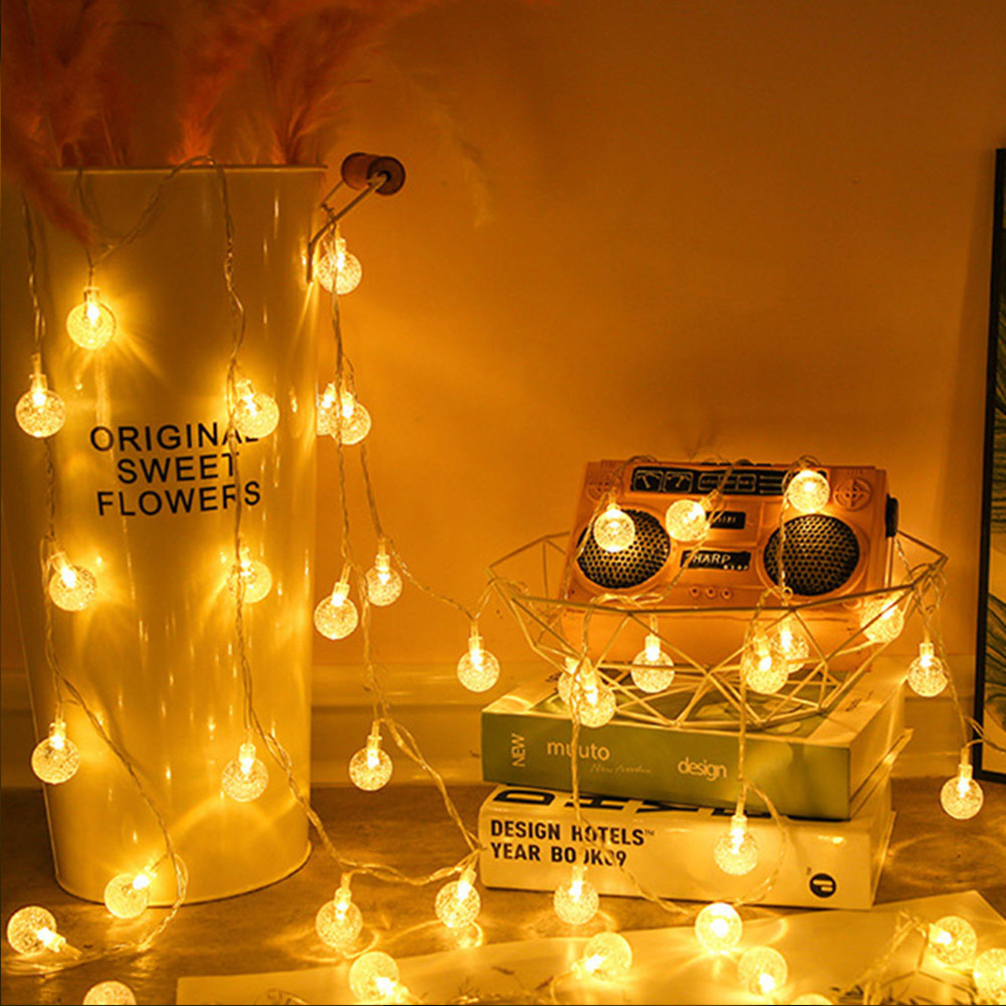 Eine Lichterkette mit Glaskugeln hängt vor einem weißen Topf mit Federn und Büchern.