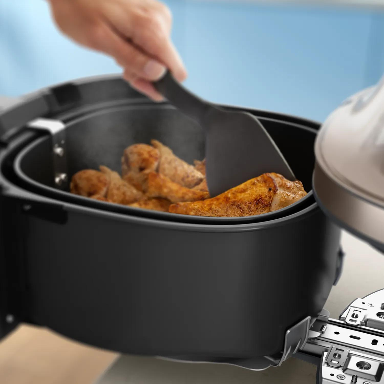 Una mano usa una spatola per girare cosce di pollo dorate in una friggitrice ad aria nera.