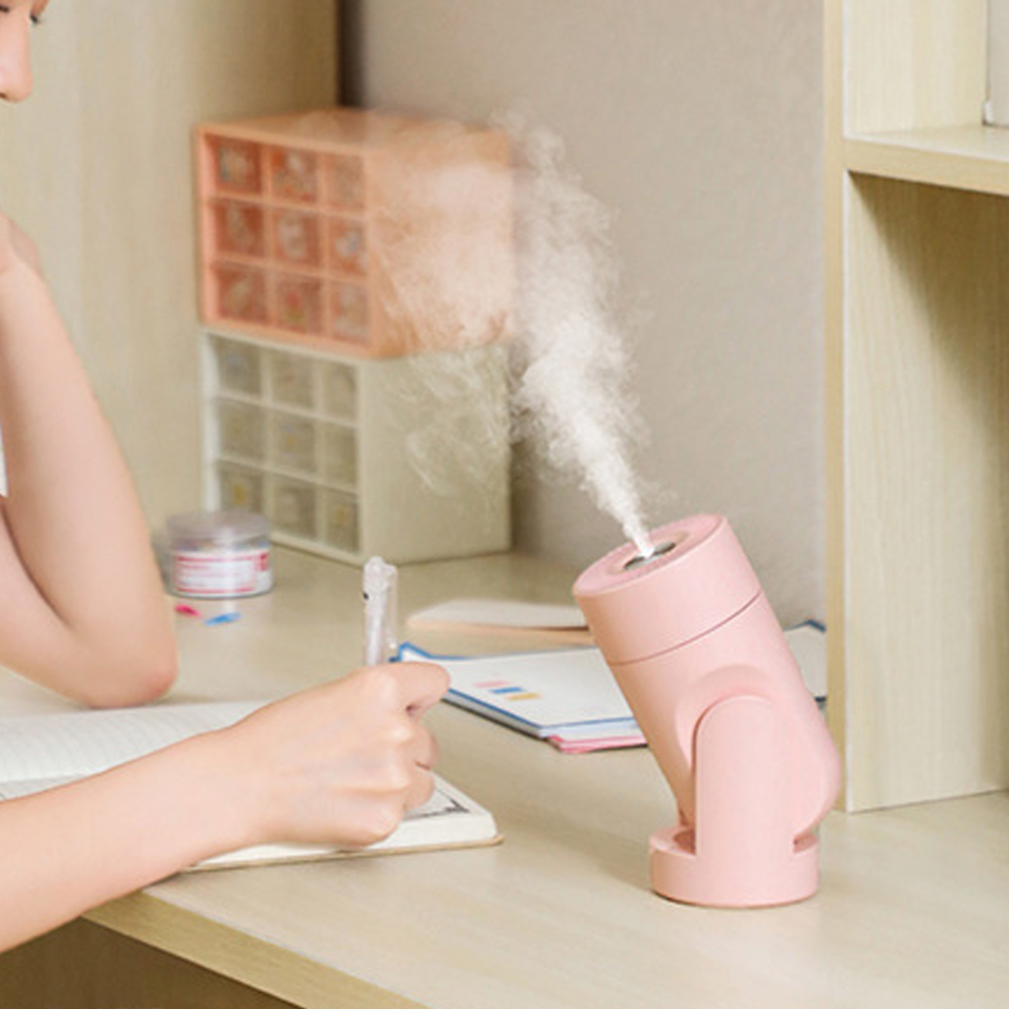 Ein rosafarbener Luftbefeuchter sprüht Nebel über einem Schreibtisch, auf dem ein Notizbuch und ein Stift liegen.